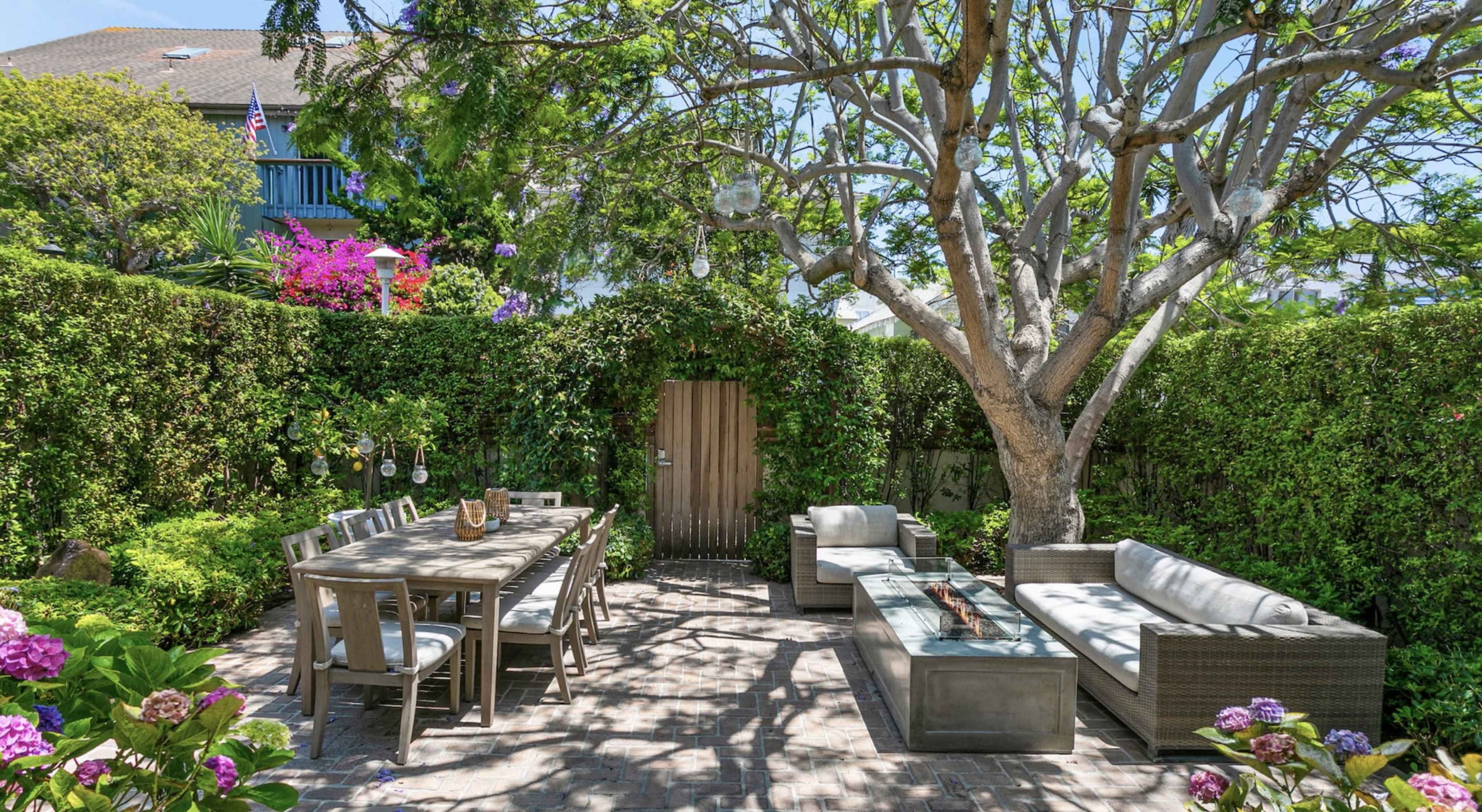 A well-furnished outdoor patio features a long dining table, comfortable seating, and lush greenery surrounding a wooden entry gate.