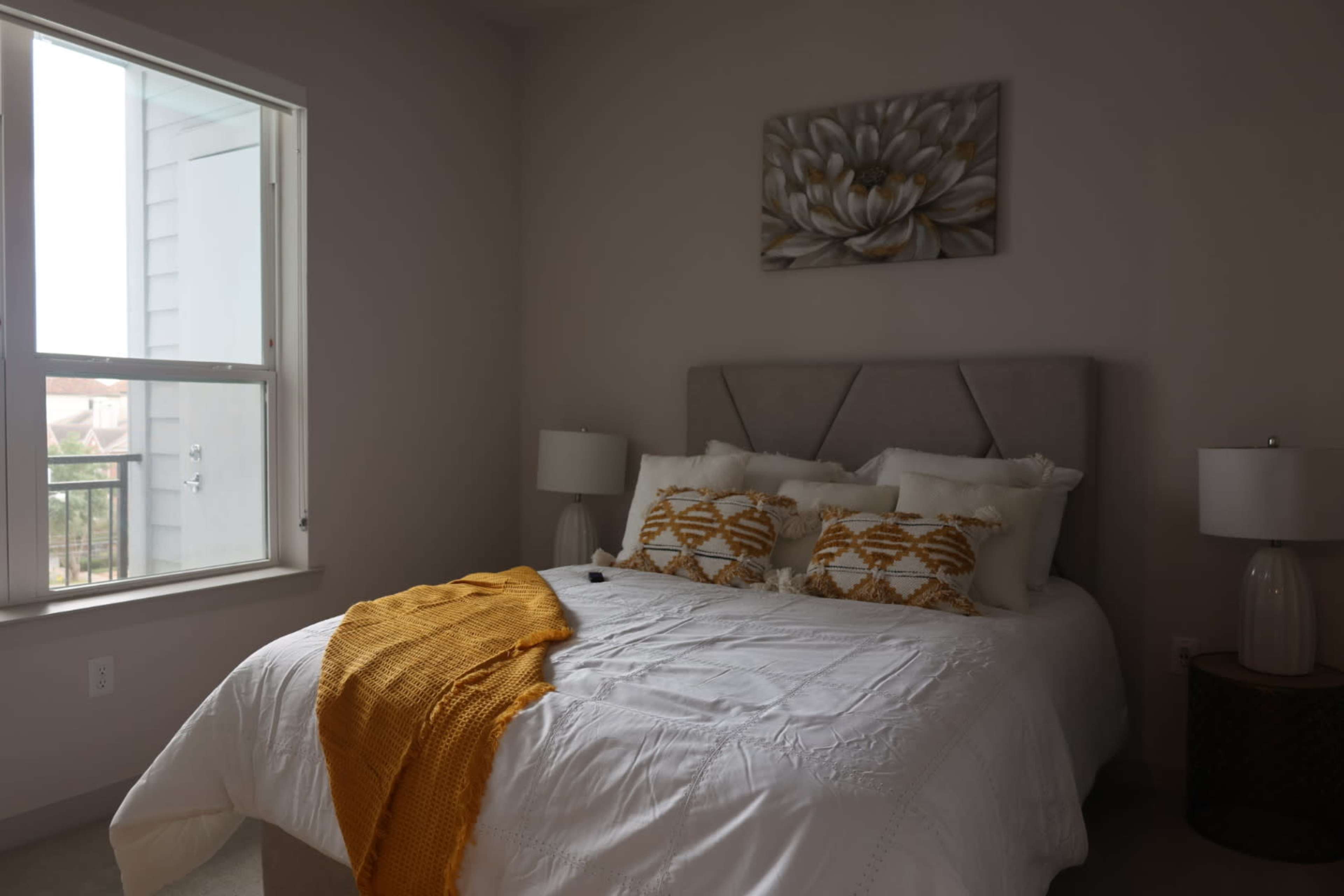 A neatly arranged bedroom features a bed with decorative pillows, a mustard-colored throw, and bedside lamps on either side.
