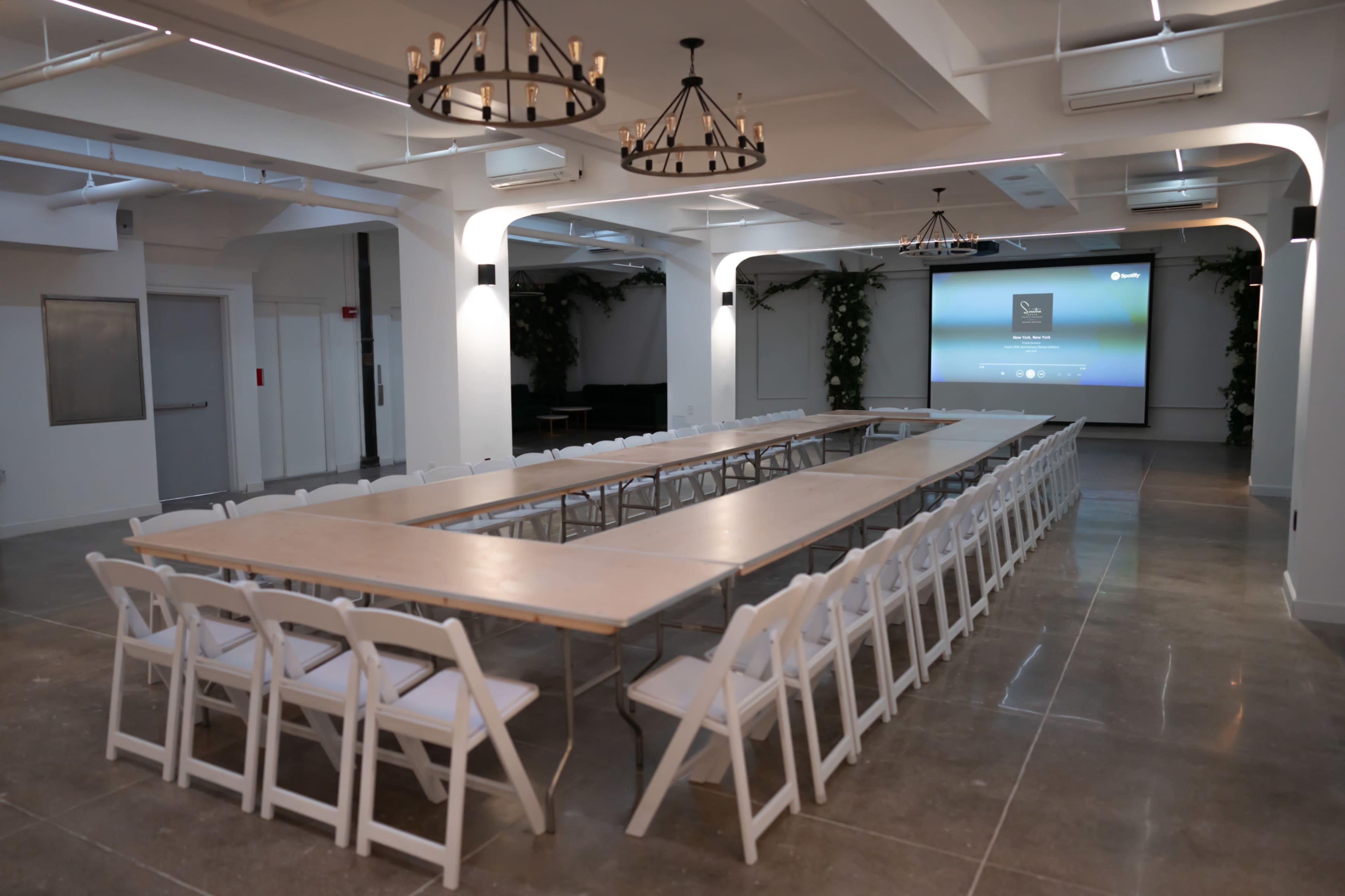 A conference room is set up with a long table and rows of white chairs, featuring a screen at the front and decorative lighting fixtures.