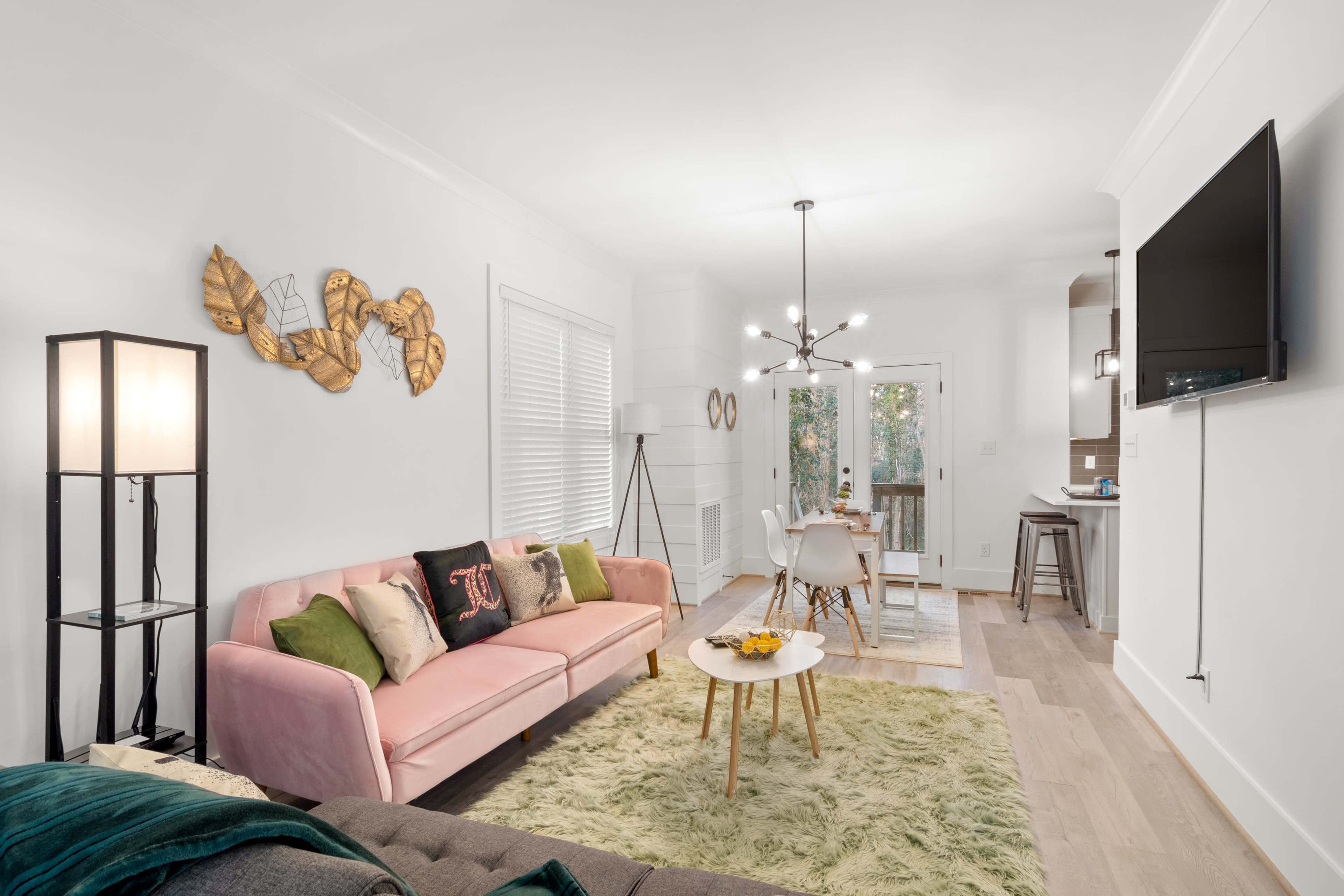A bright living room featuring a pink sofa, a wooden coffee table with fruit, and a dining area visible in the background.
