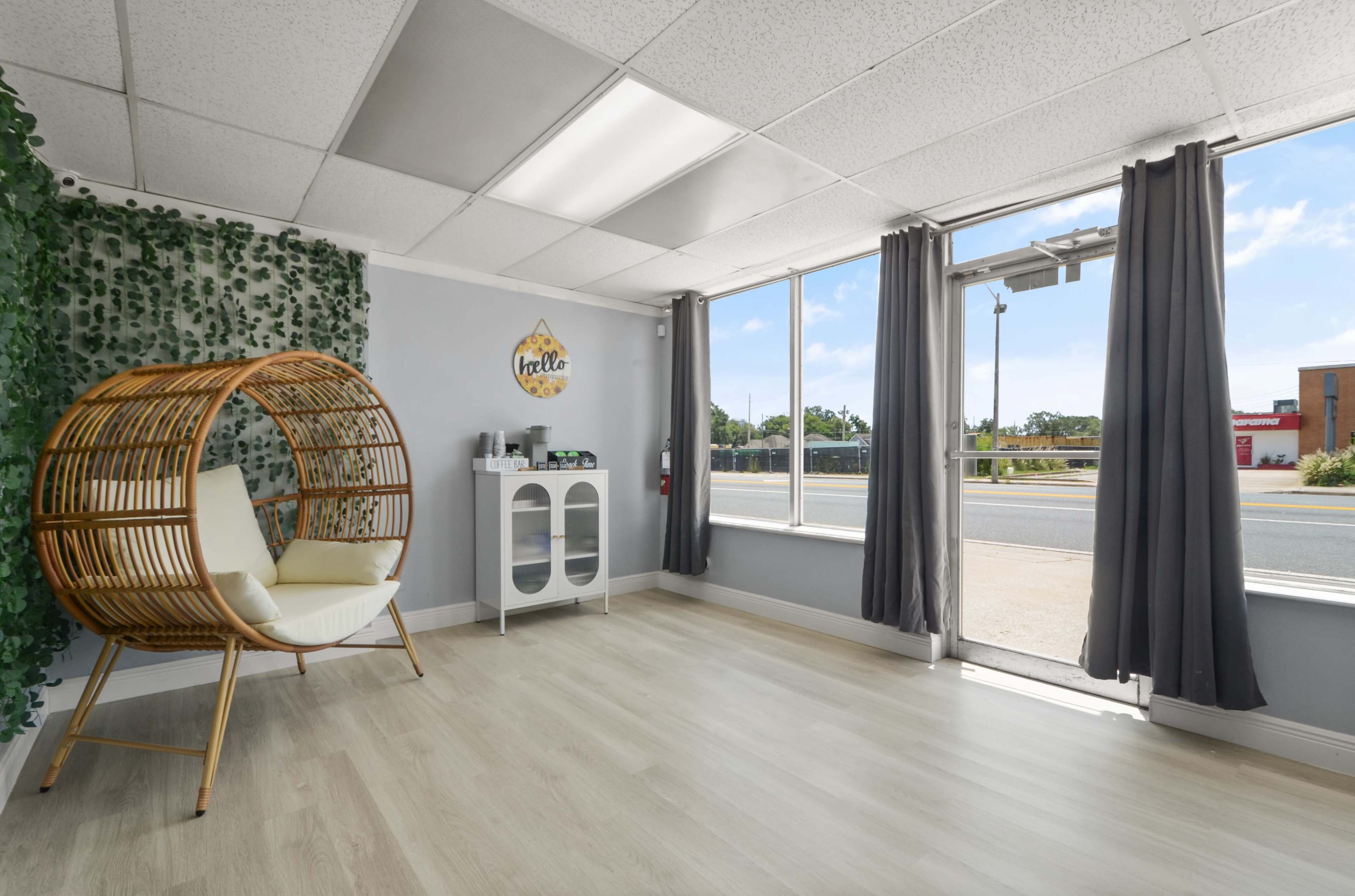 The image shows a bright, open room with a rattan chair, a small cabinet, and large windows that provide a view of the outside.