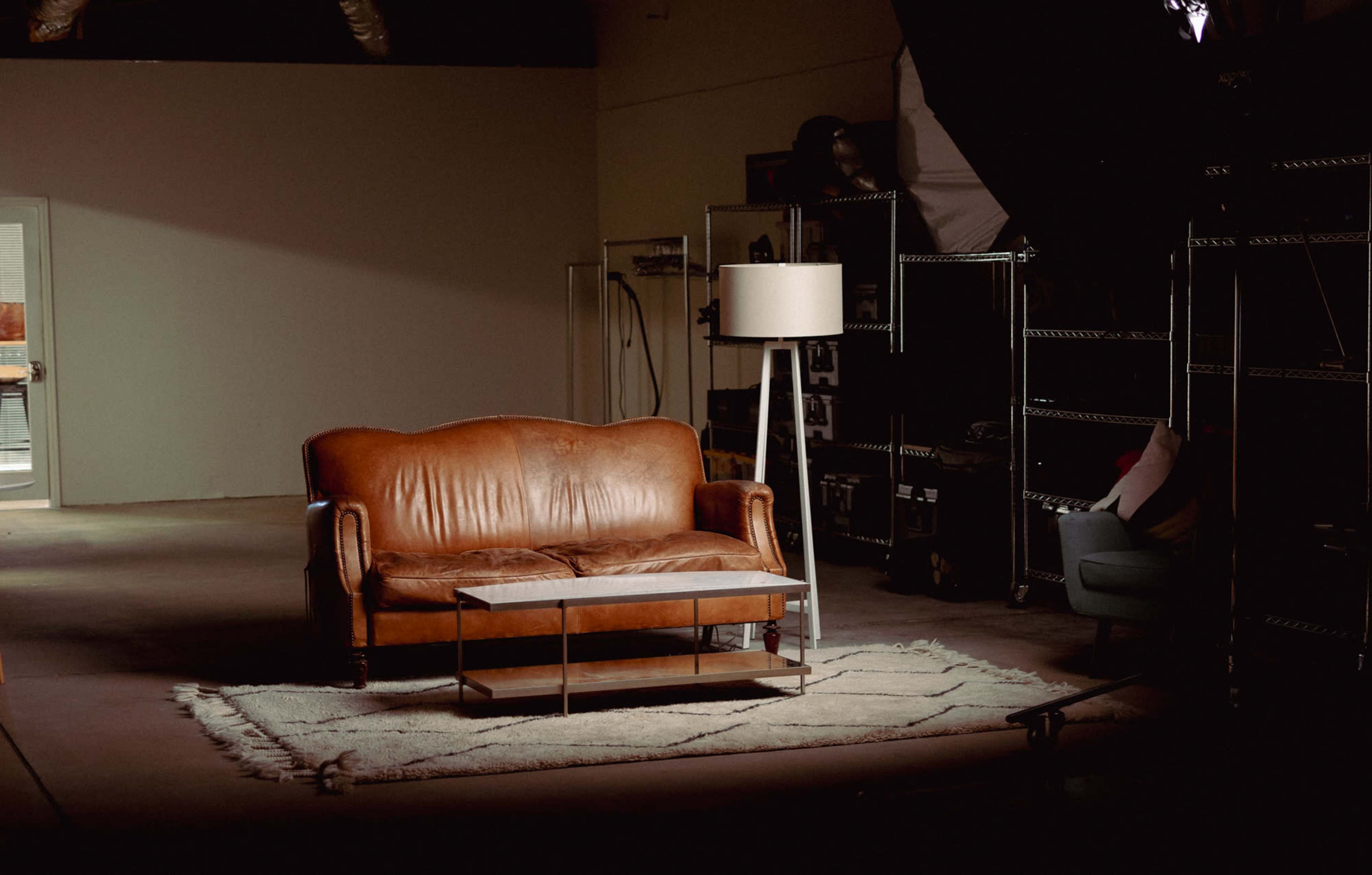 A brown leather sofa is positioned beside a small table on a textured rug in a dimly lit studio space, with storage shelves in the background.