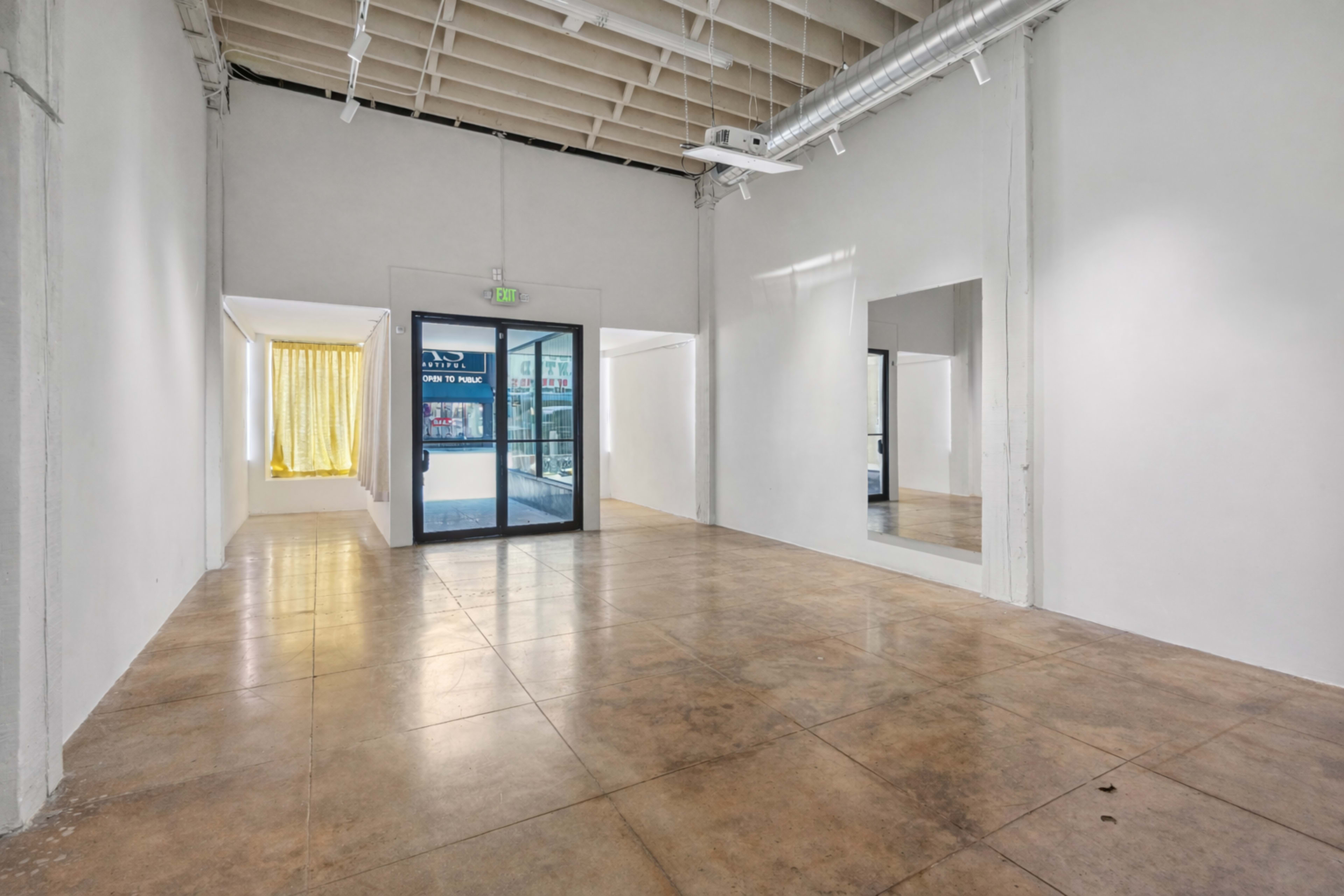 The image shows a spacious, empty interior room with concrete floors and large windows providing natural light.