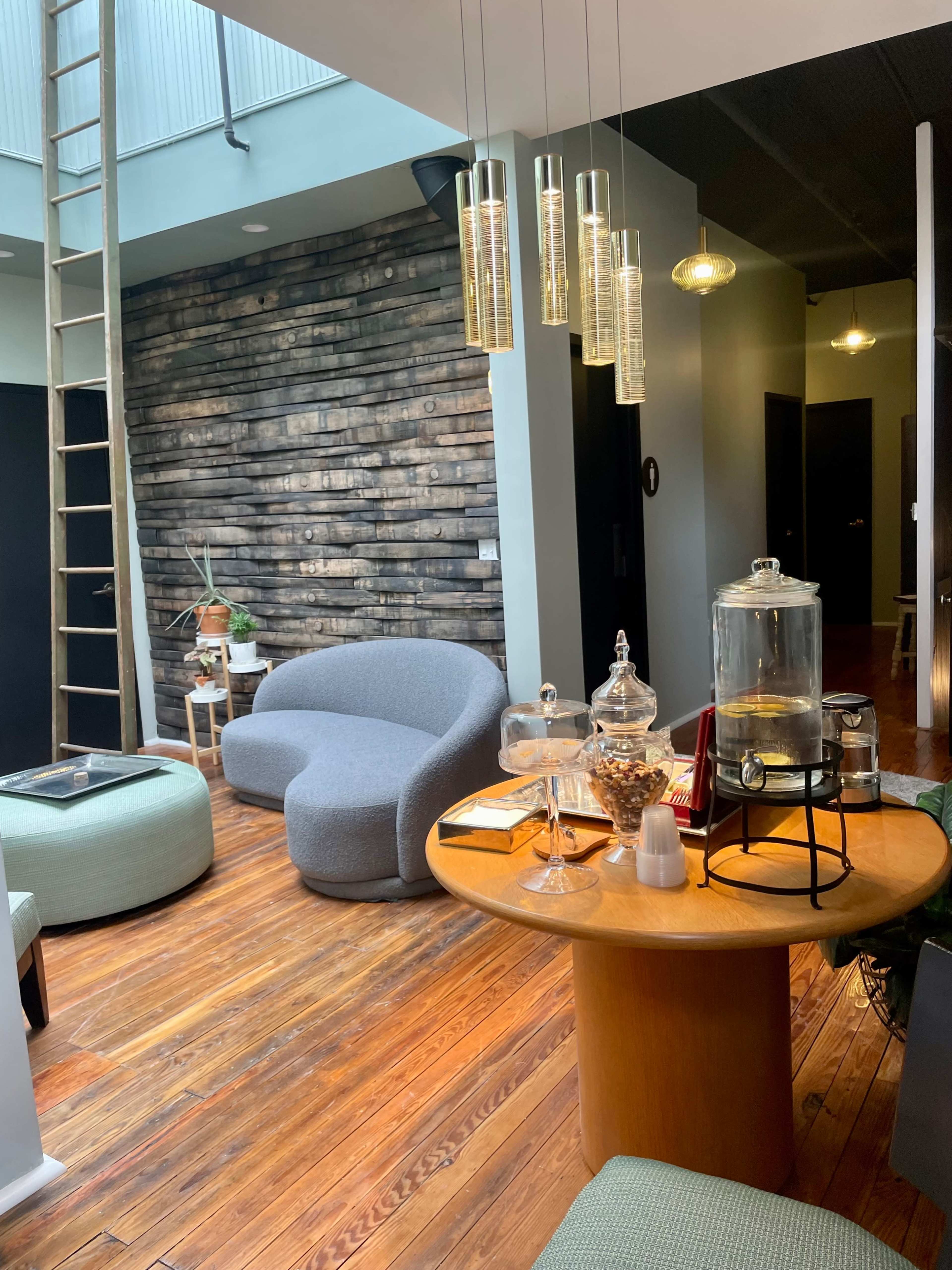 A modern interior with a wooden floor, a round table displaying a glass jar, and a cozy seating area featuring a curved sofa and poufs.