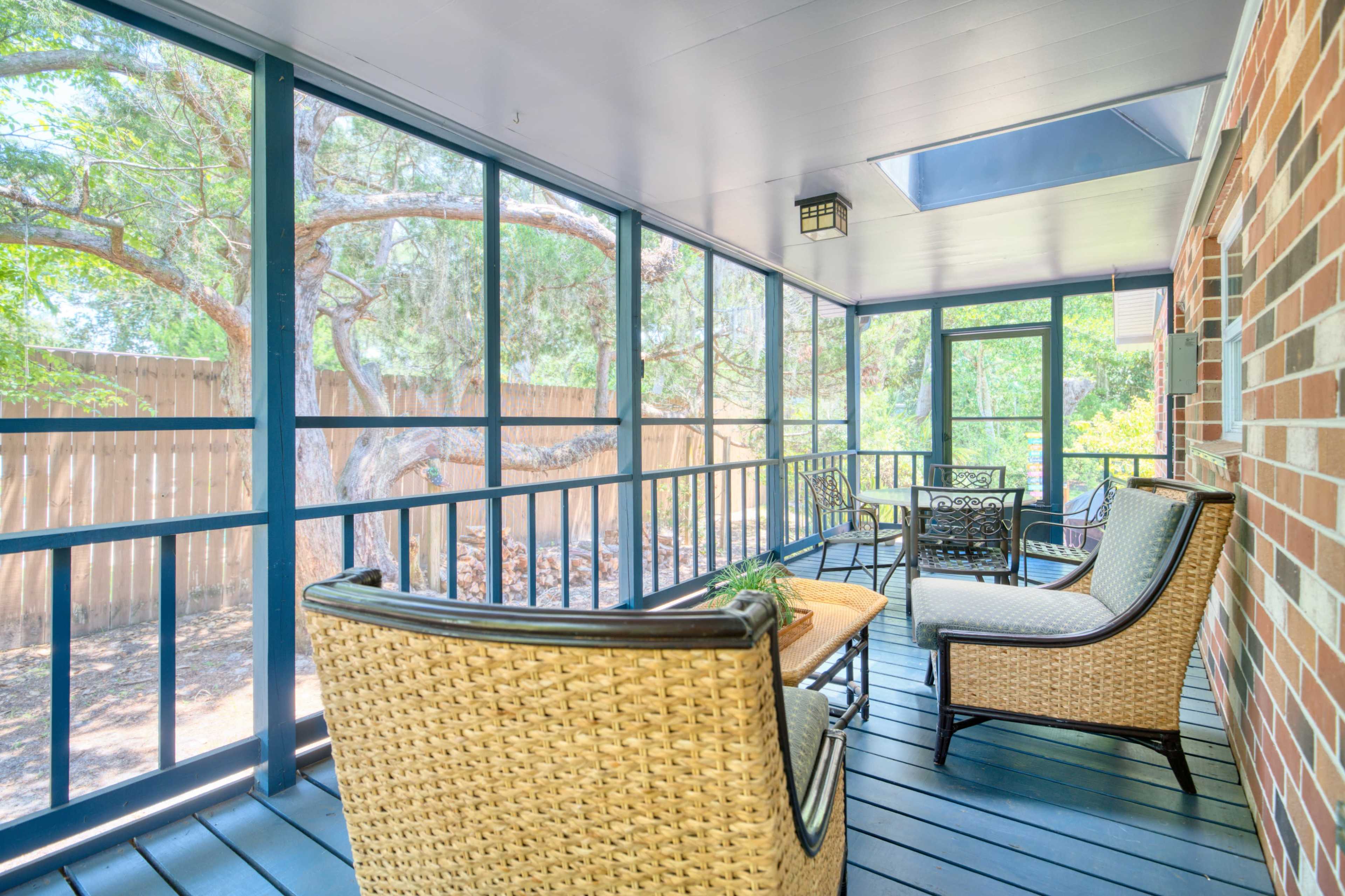 A screened porch with wicker furniture, including two chairs and a small table, overlooking a backyard with trees.