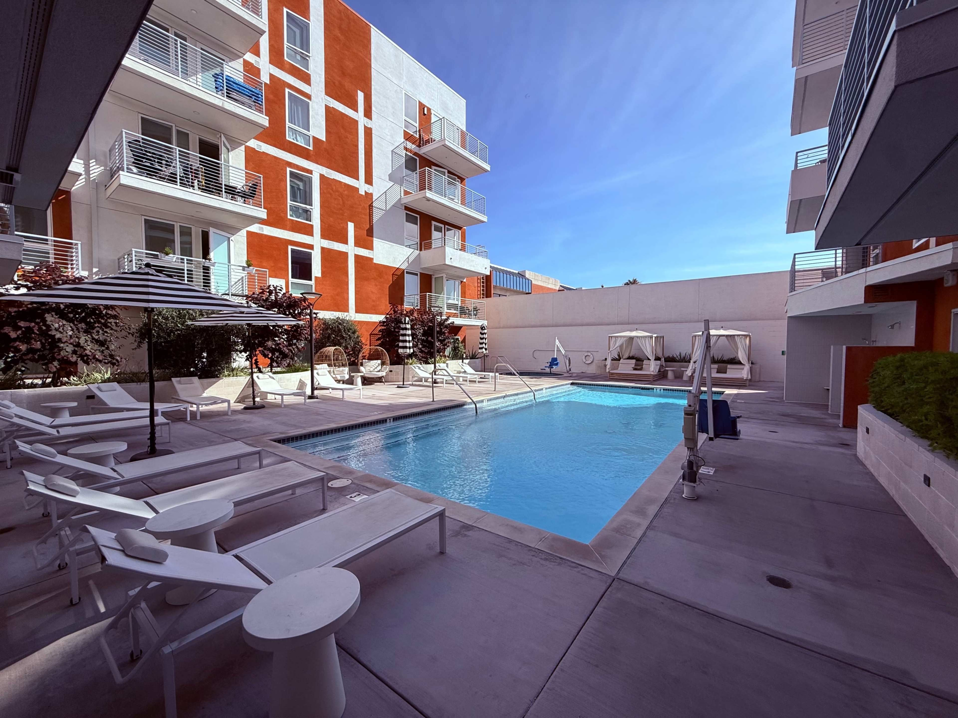 The image shows a modern pool area surrounded by multi-story apartment buildings, with lounge chairs and cabanas arranged around the pool.