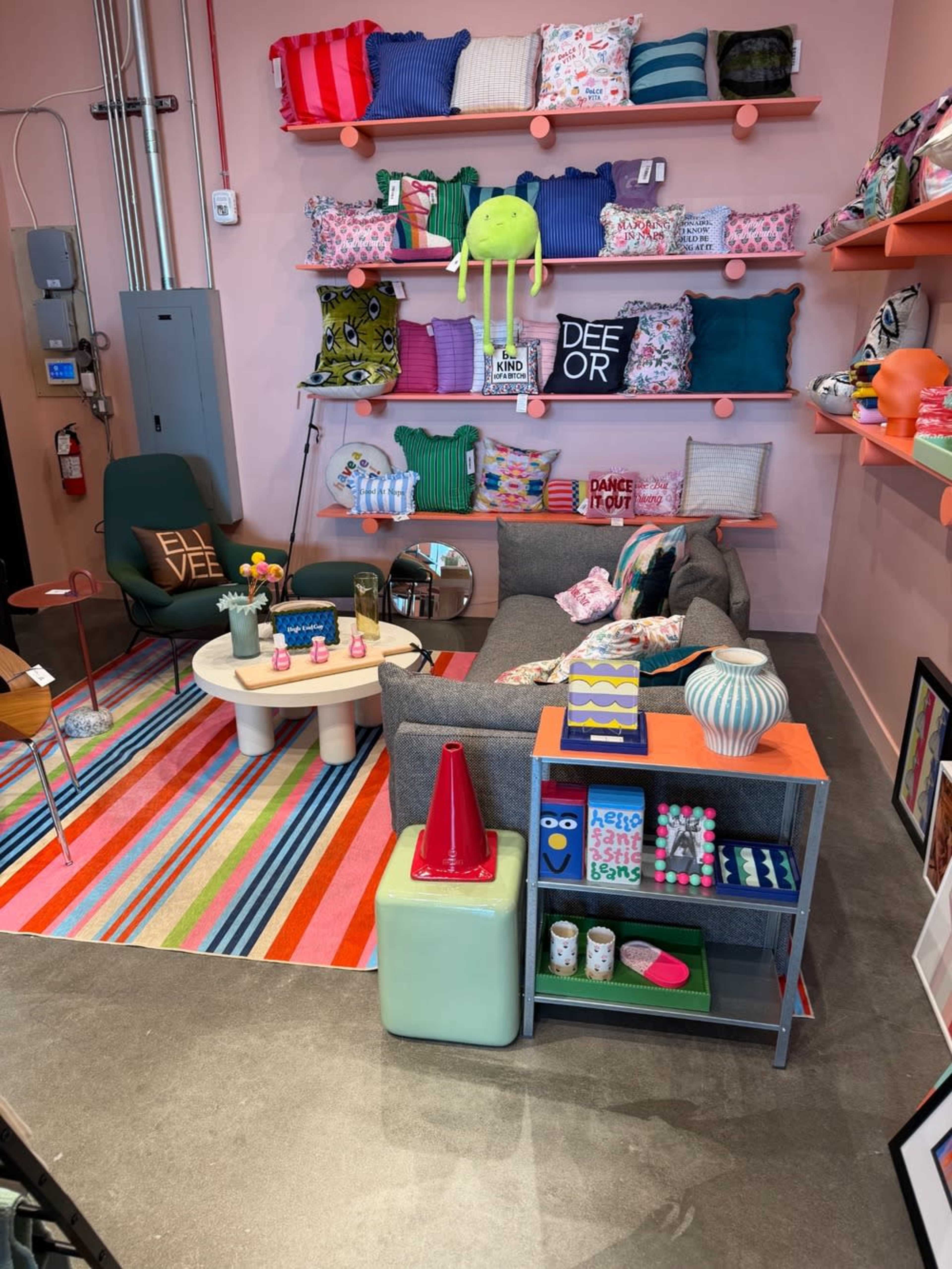 The image shows a colorful interior scene featuring a cozy seating area with a gray sofa, a round coffee table, and various decorative items on shelves and a cart, against a light pink wall.