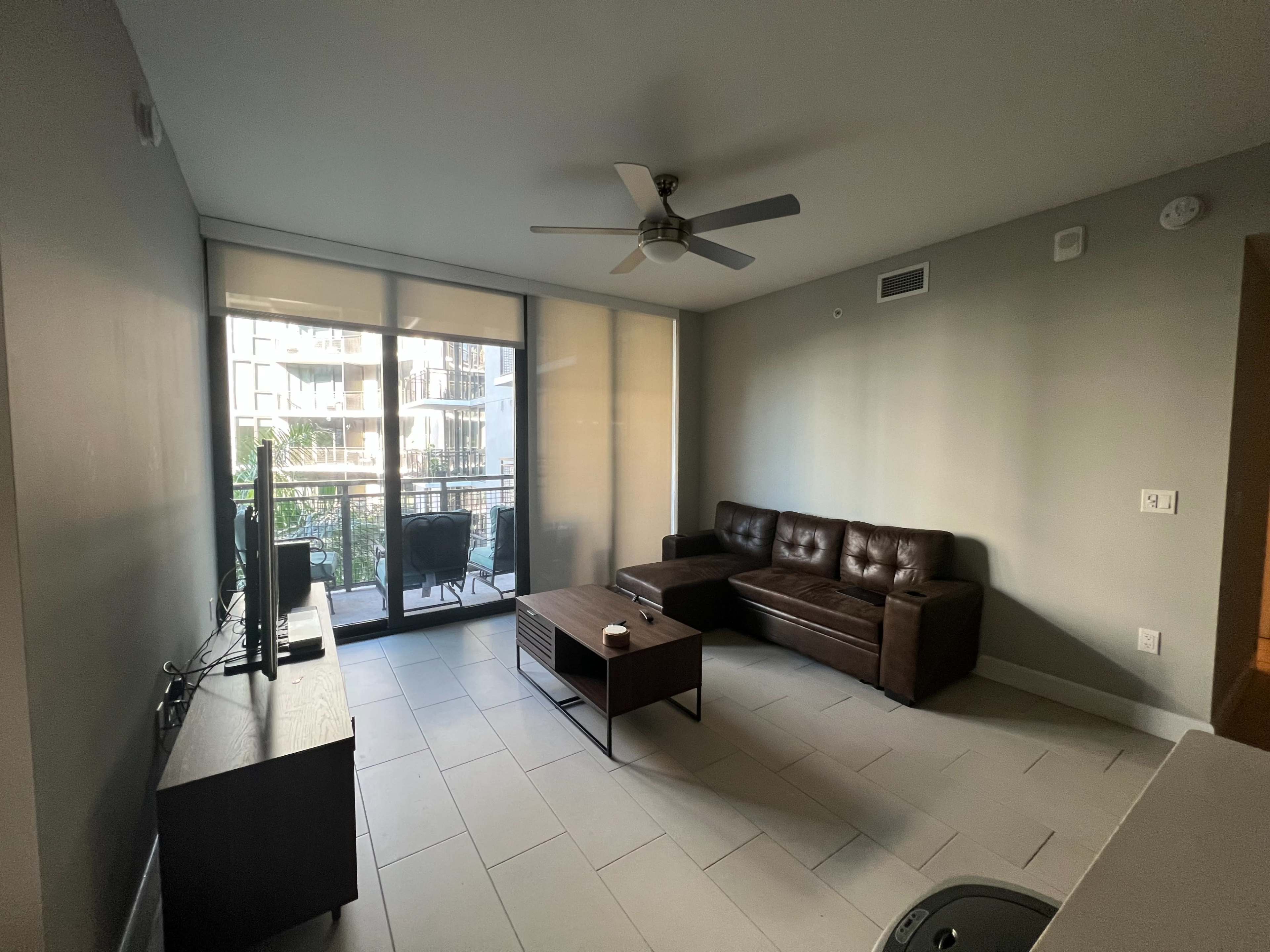A modern living room with a brown sofa, a coffee table, a television, and large windows overlooking a balcony.