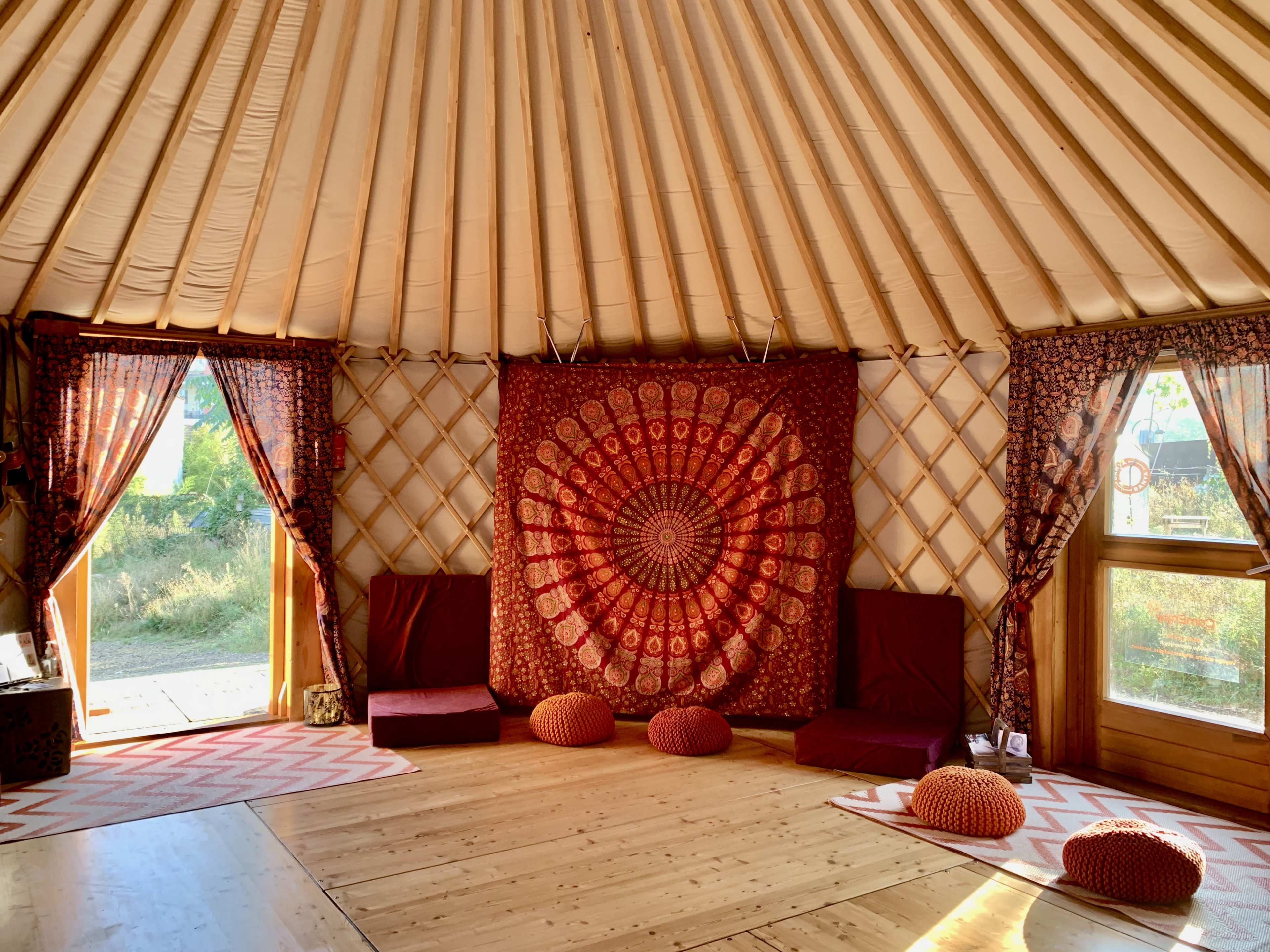 The interior of a yurt features a large circular tapestry, floor cushions, and wooden floors, with light coming through two windows adorned with patterned curtains.