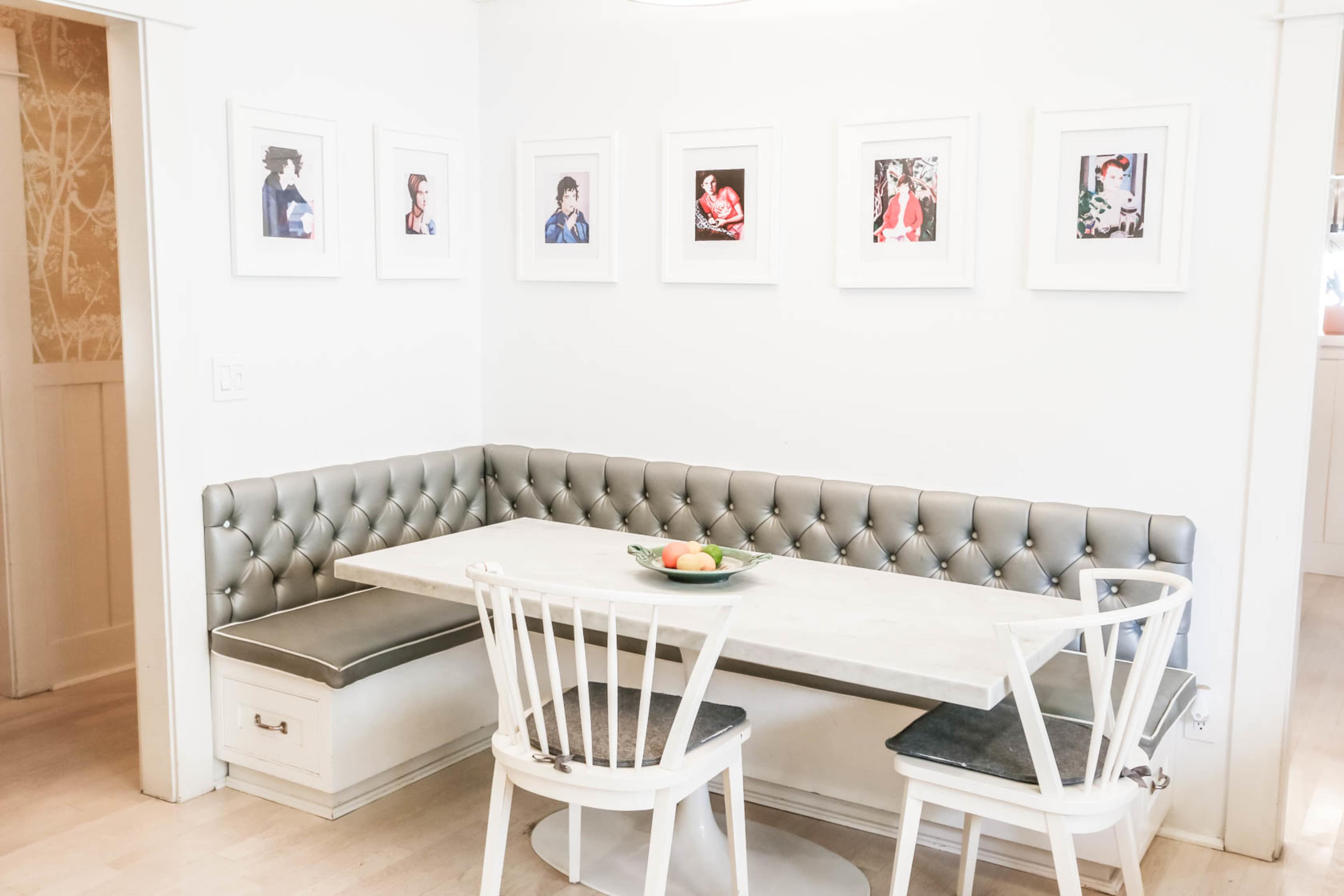 A dining area features a tufted gray bench along the wall, a rectangular white table, and two white chairs, with framed artwork displayed above.
