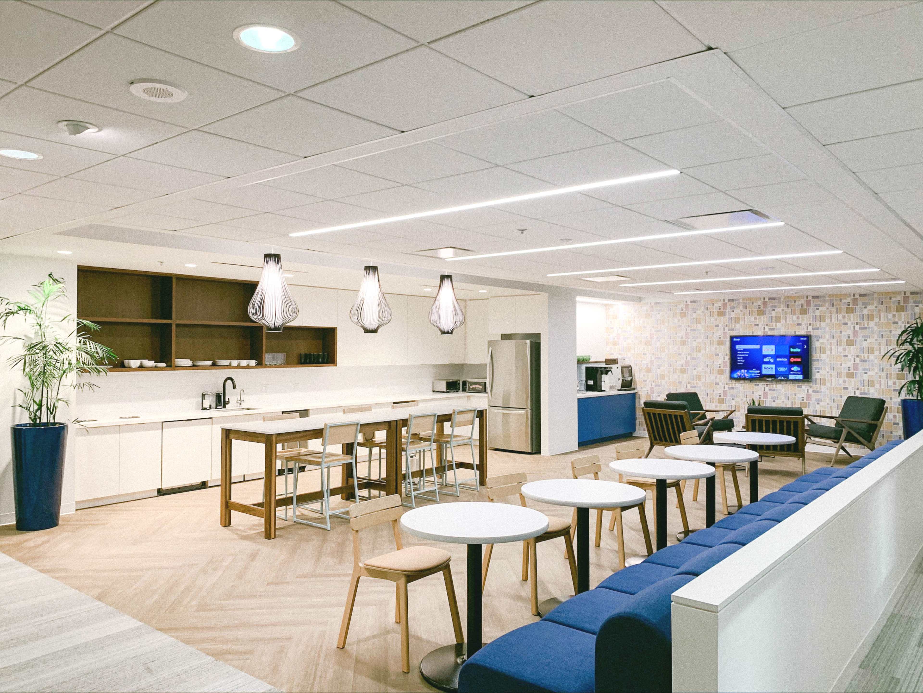 A modern office kitchen area with a dining table, chairs, light fixtures, and a coffee machine against a background of patterned wallpaper and a television.