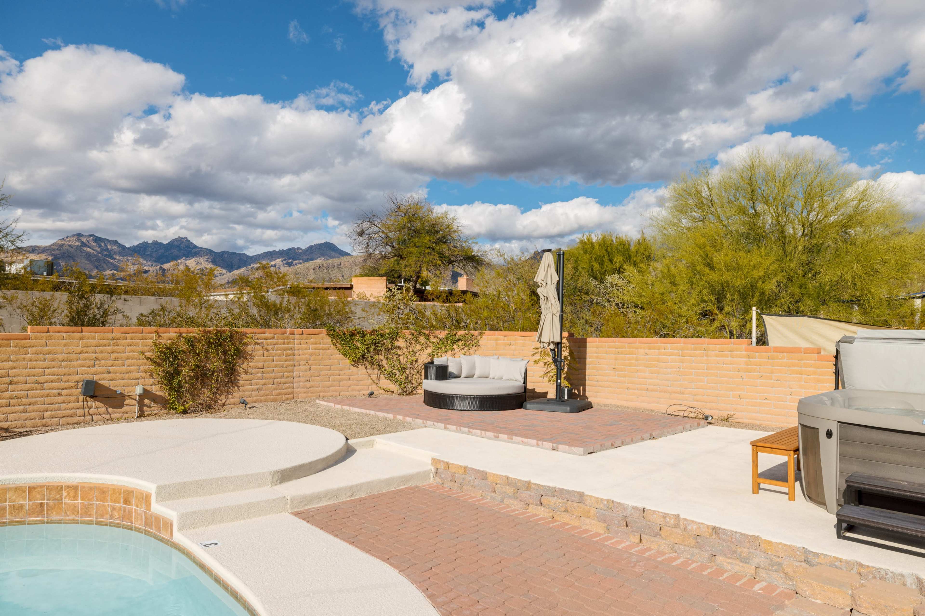 A backyard patio with a hot tub, a circular seating area, and landscaped stone and concrete surfaces against a backdrop of mountains and clouds.