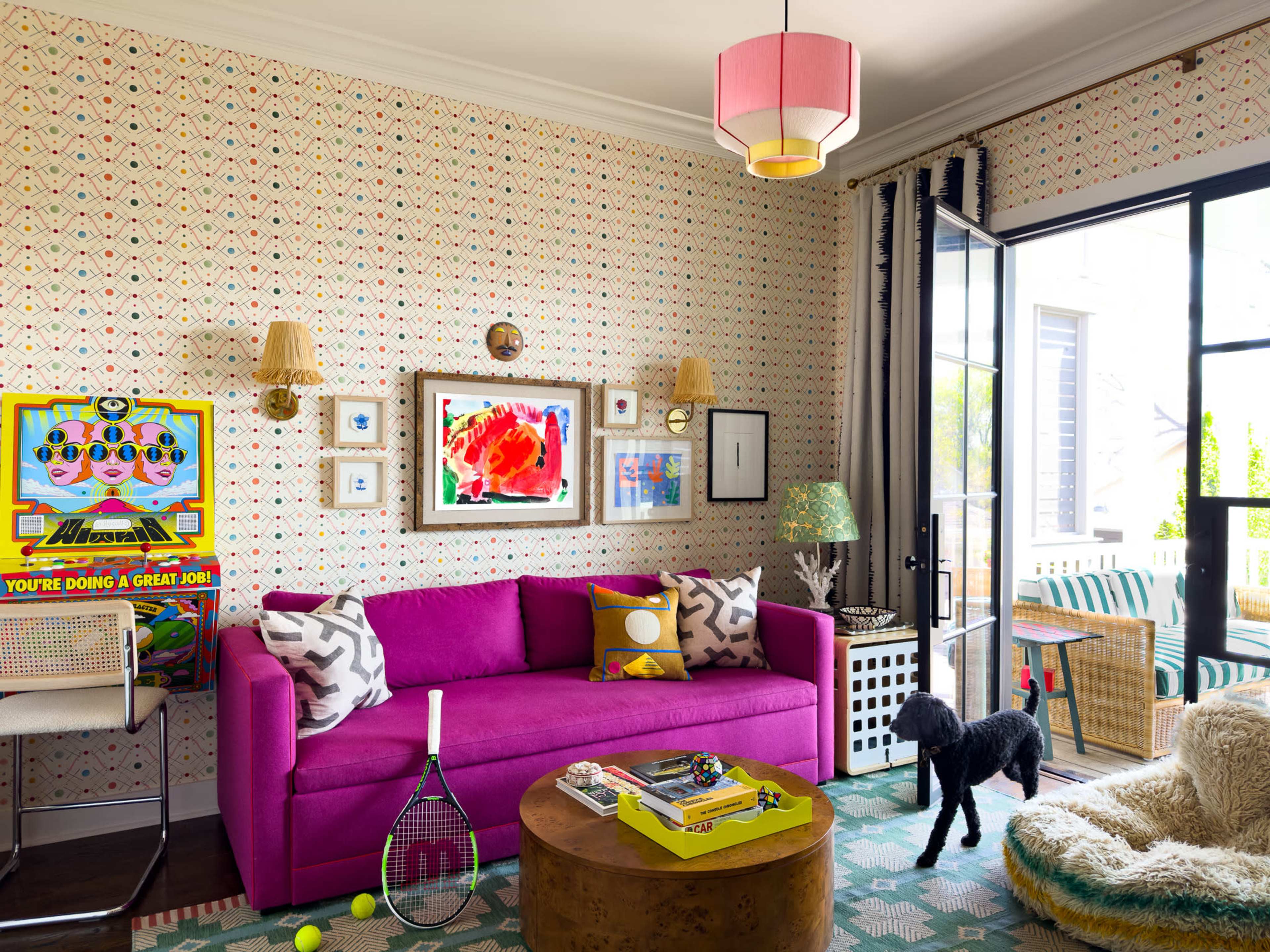 The room features a vibrant pink sofa, a round coffee table, and various pieces of wall art, with a tennis racket and balls on the floor alongside a black dog.