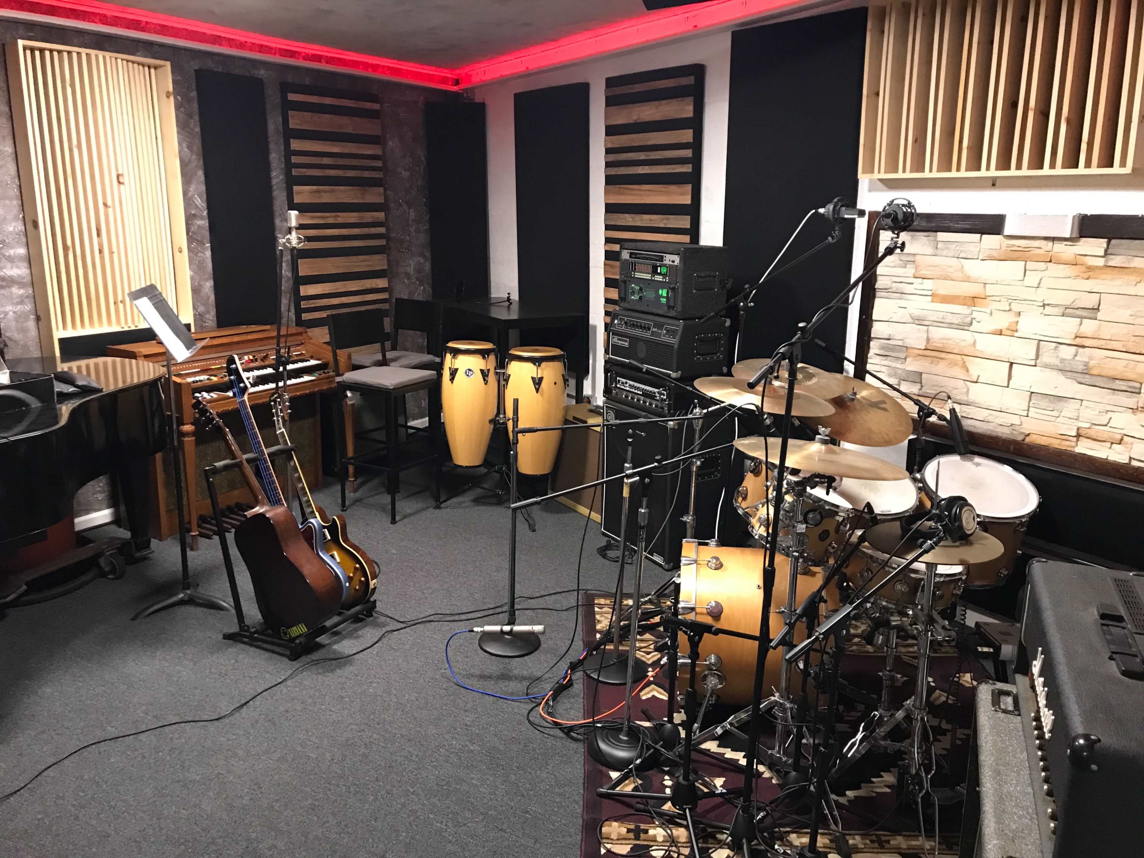 The image shows a music studio with various instruments, including a drum set, congas, an electric guitar on a stand, and a grand piano, surrounded by sound equipment and soundproofing materials.