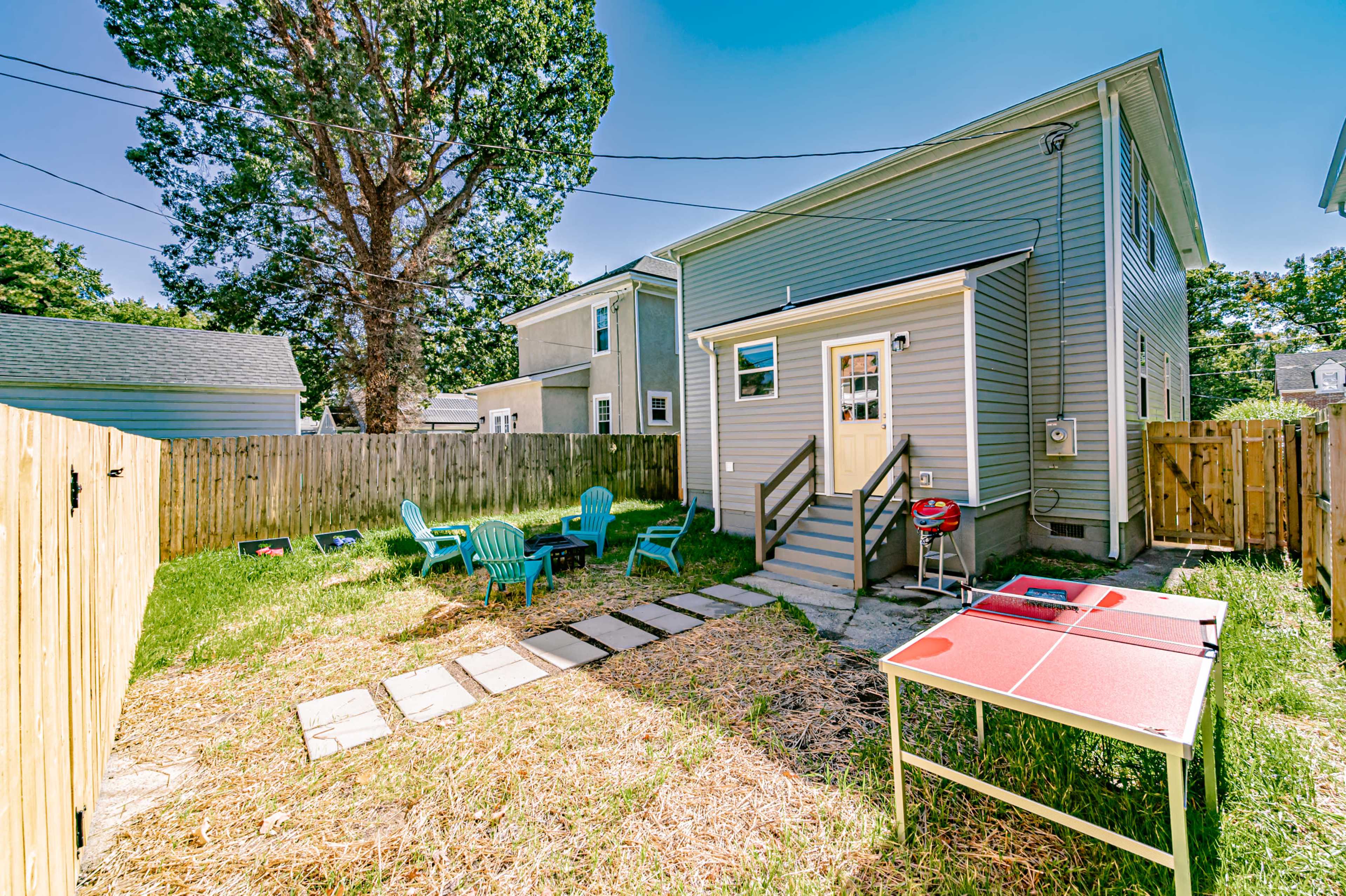 A backyard features a small patio area with green chairs, a barbecue grill, a ping pong table, and a pathway made of stone slabs.