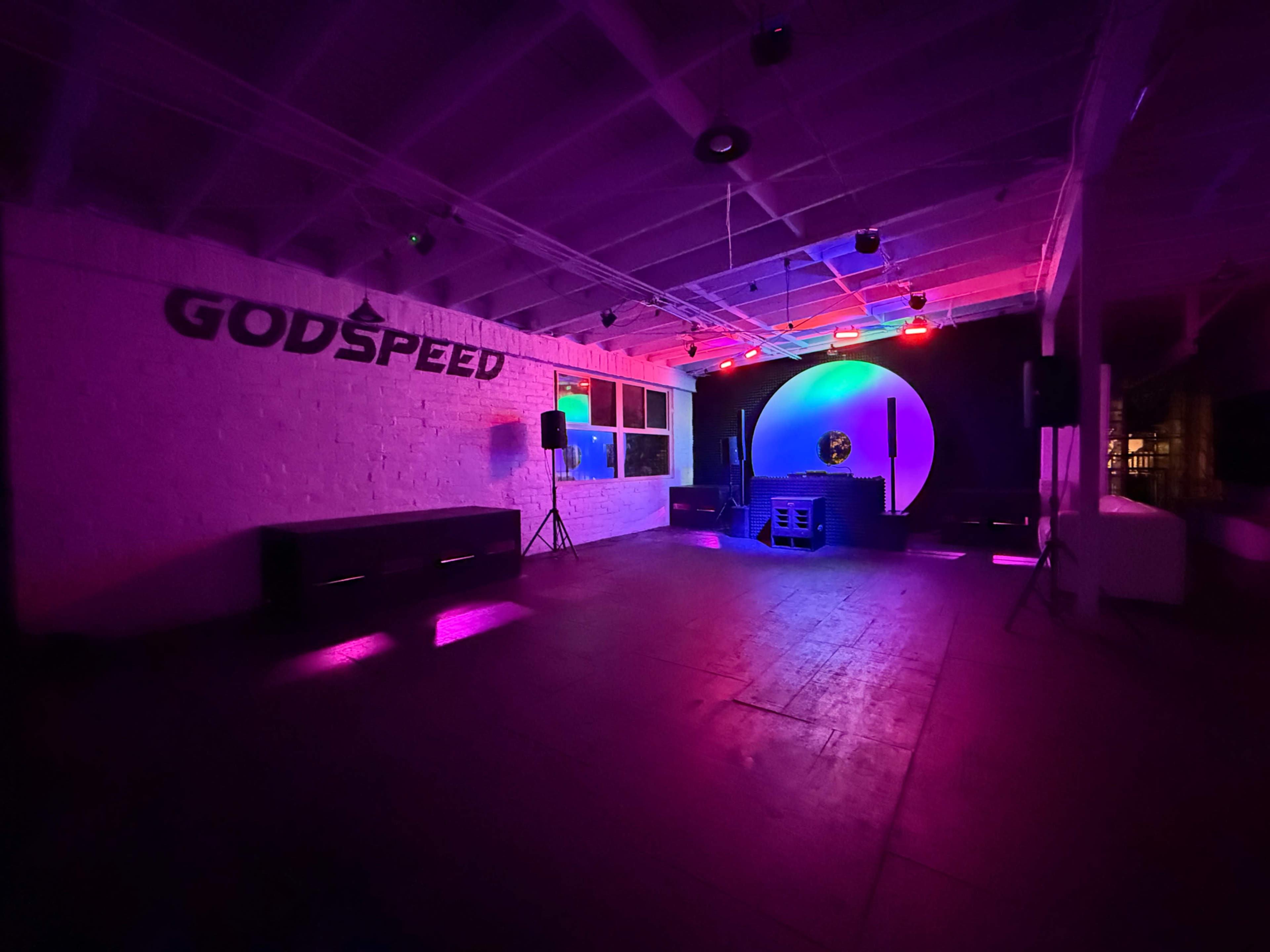 A dimly lit event space features a large round backdrop with colored lights, the word "GODSPEED" on one wall, and sound equipment set up for a performance.