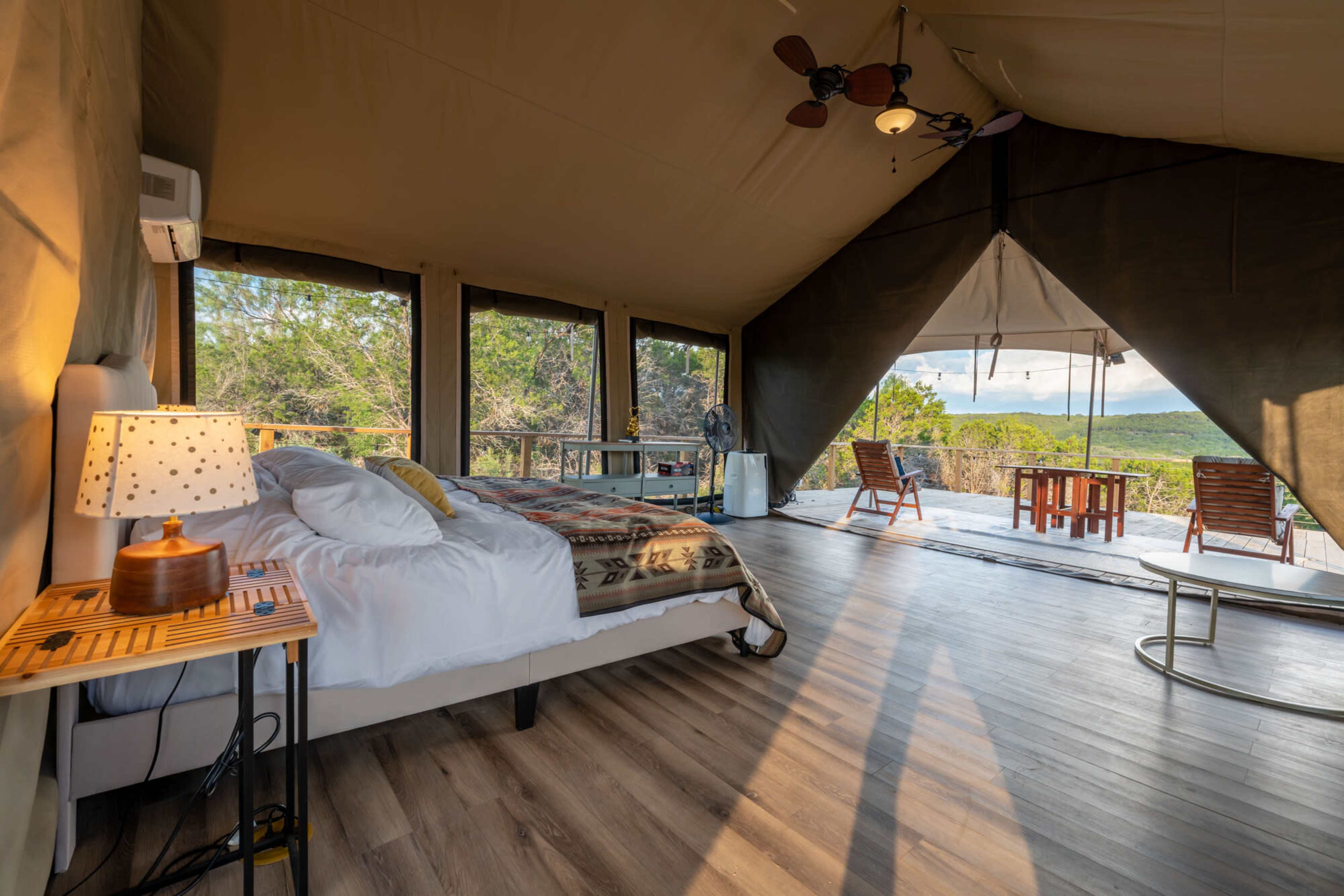 The image shows a spacious glamping tent interior featuring a bed, table, and chairs with large windows that provide a view of the outdoors.