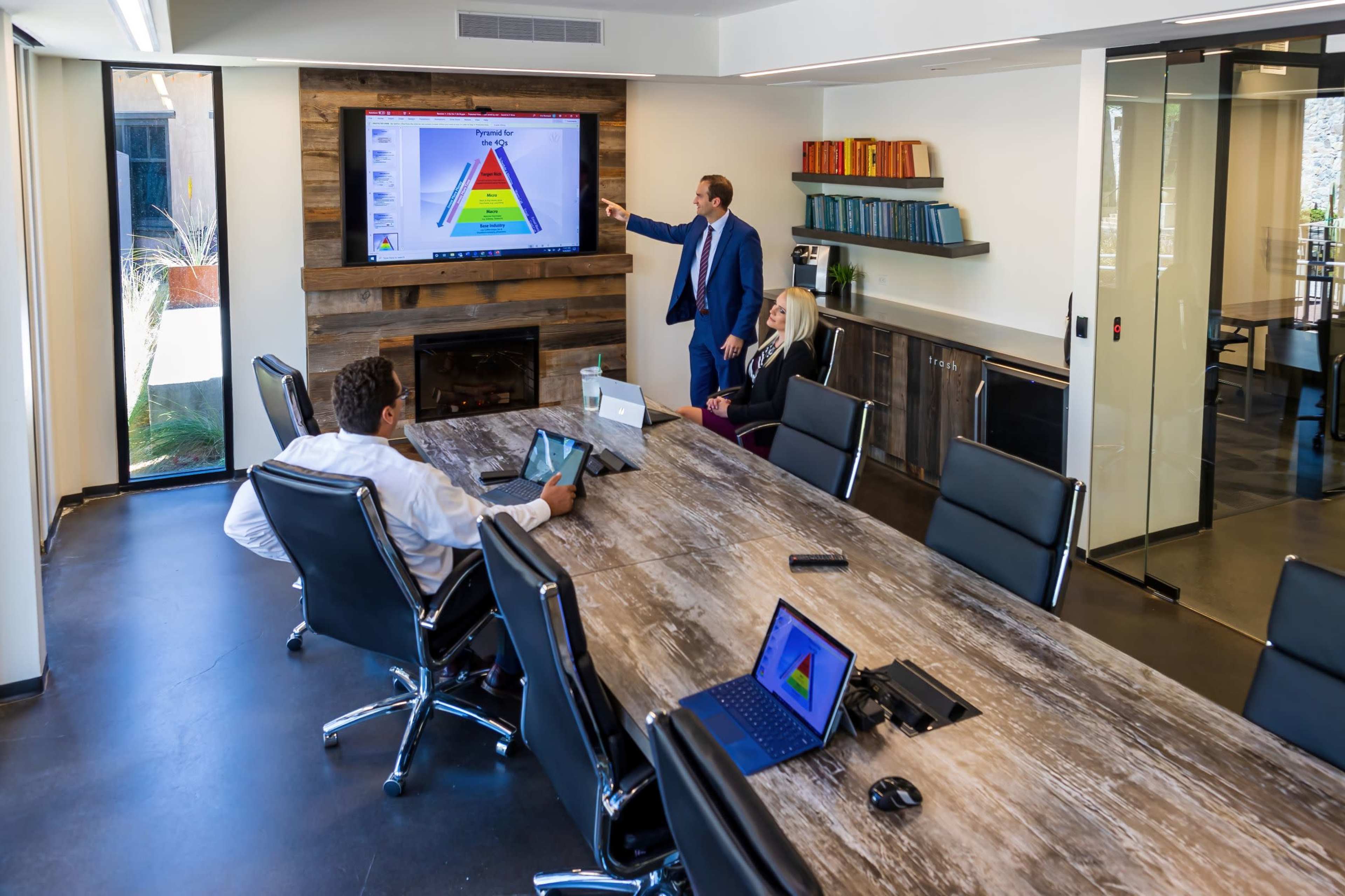 A group of professionals is engaged in a presentation in a modern conference room, with one person pointing at a screen displaying a colorful pyramid diagram.