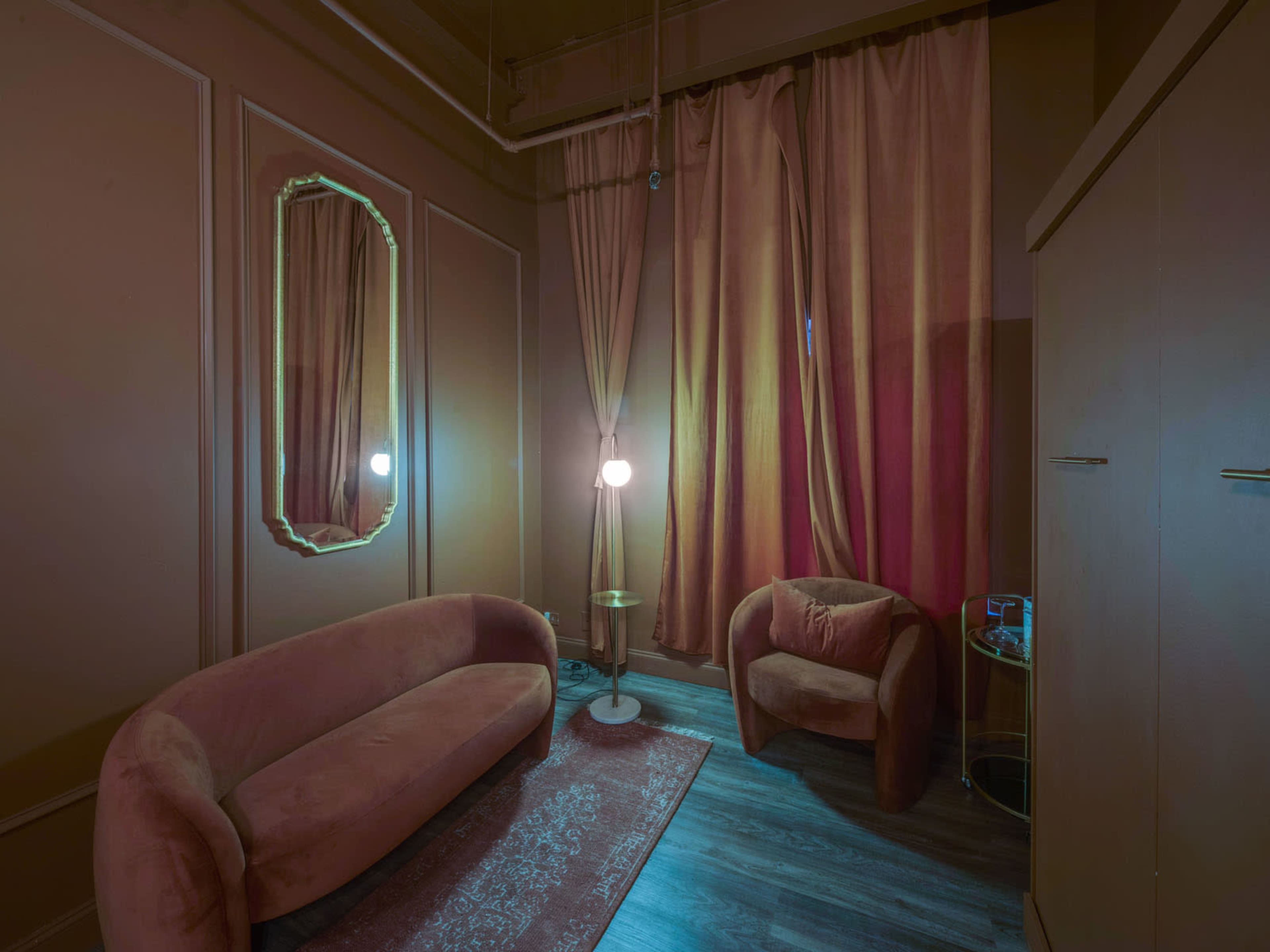 A small, dimly lit room featuring a curved sofa, an armchair, a mirror, a floor lamp, and curtains, all in warm tones.