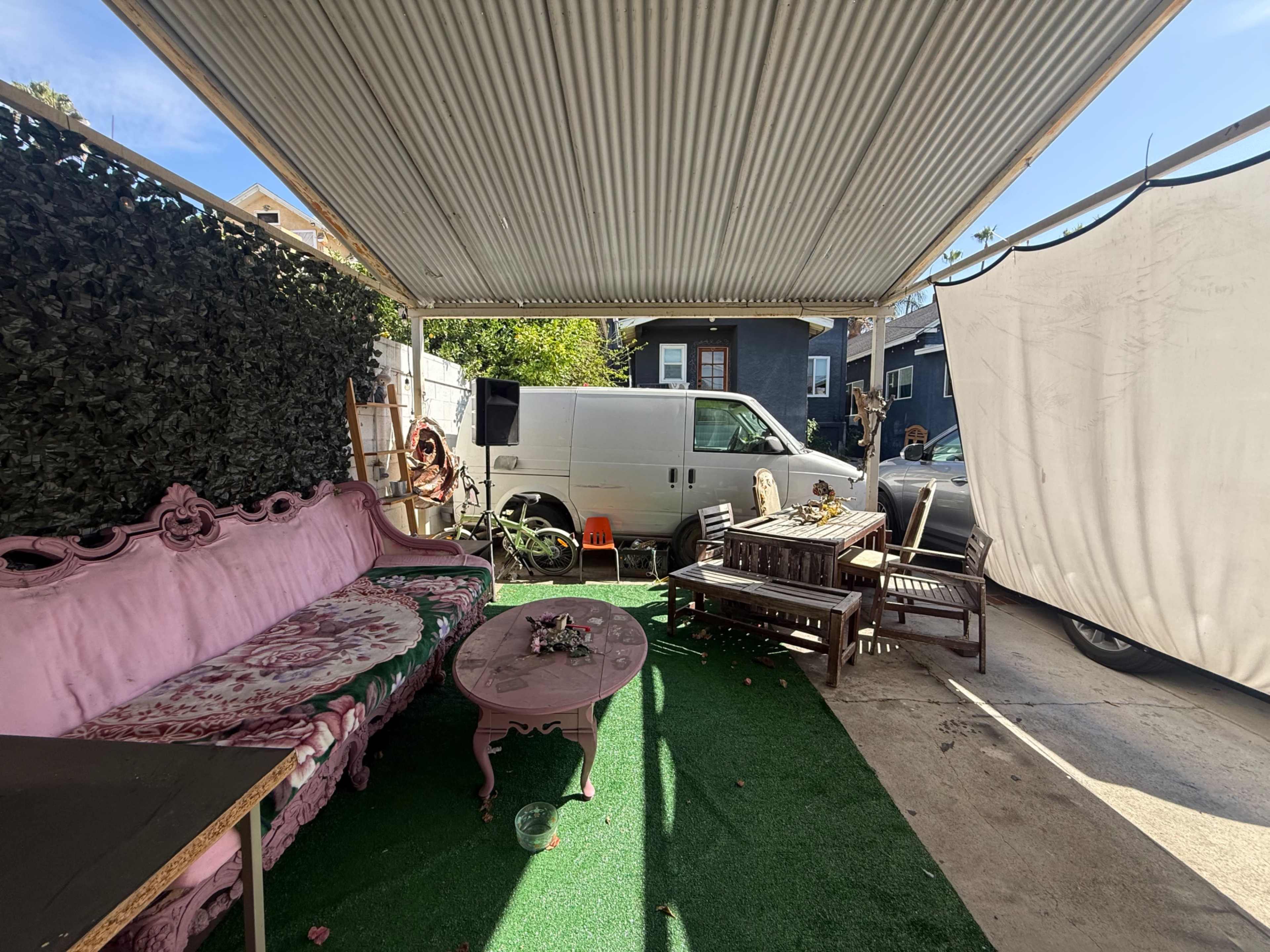 The image shows a covered outdoor area with pink furniture, including a couch and a table, next to a white van and a wall of greenery.