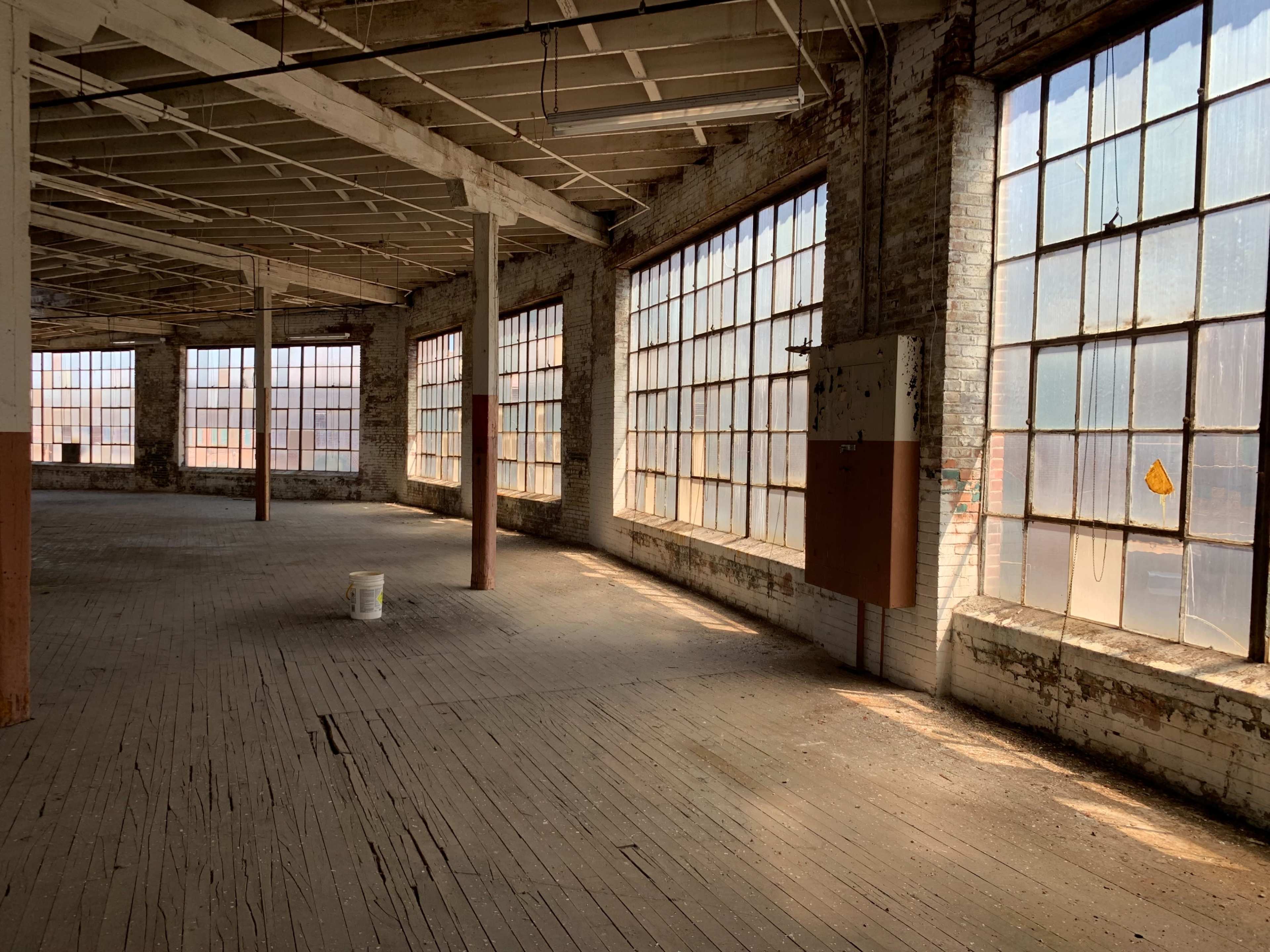 The image shows an empty, large warehouse with multiple tall windows allowing light to filter in, highlighting the wooden floor and brick walls.