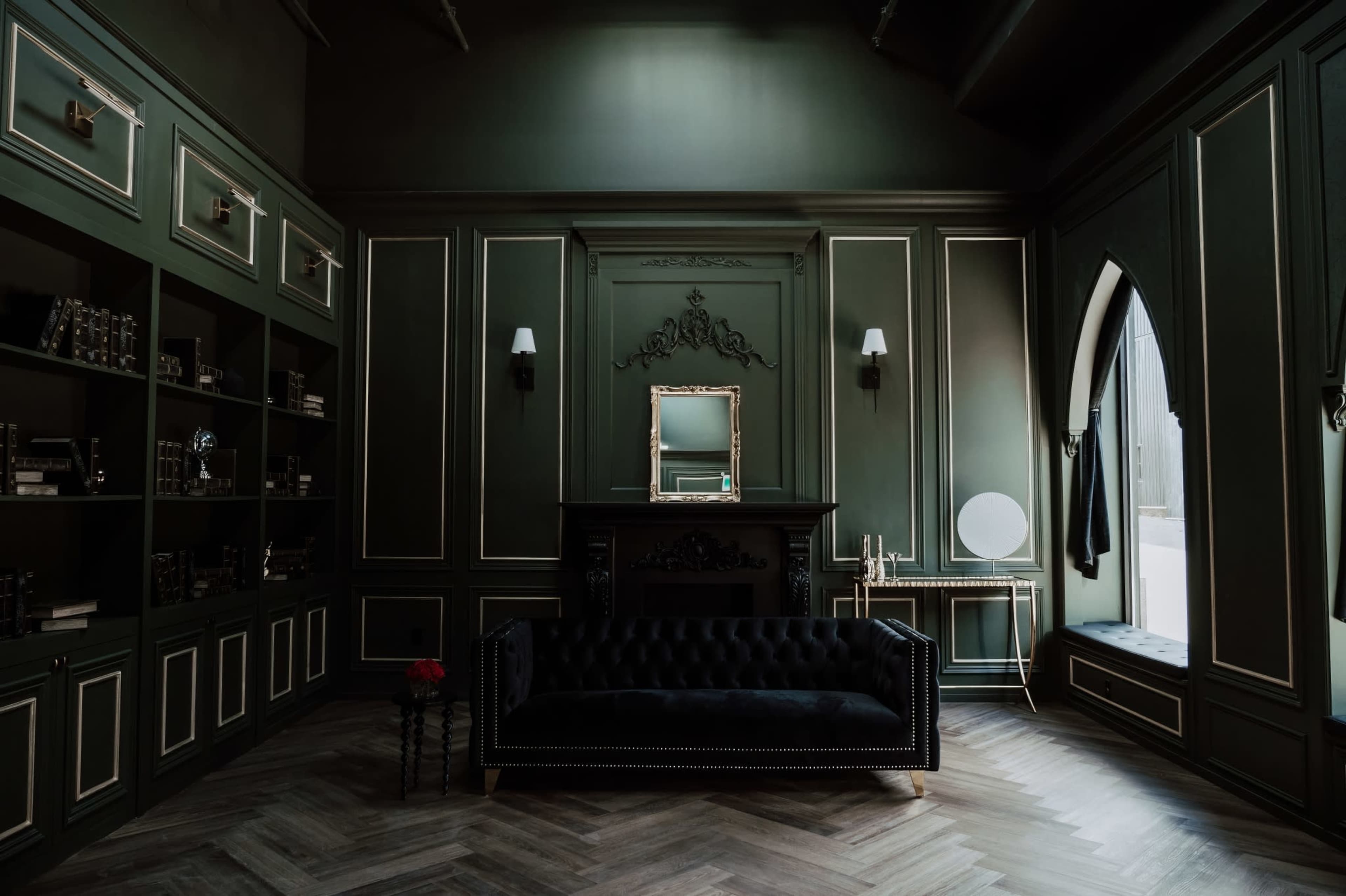 The room features dark green walls with ornate trim, a black velvet sofa, and a large window alongside bookshelves and decorative accents.