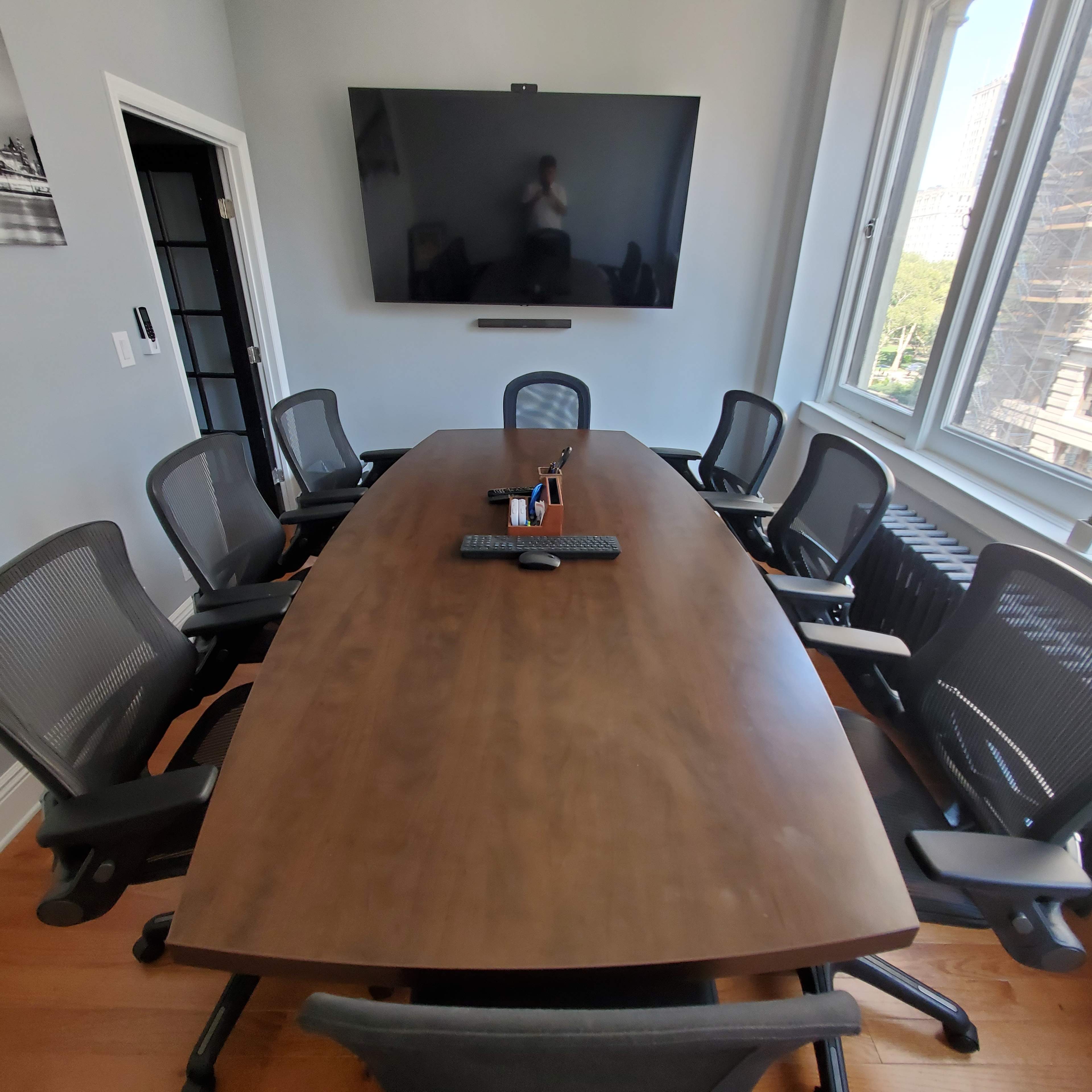 A large, oval wooden conference table is surrounded by ergonomic chairs in a brightly lit meeting room featuring a wall-mounted screen.