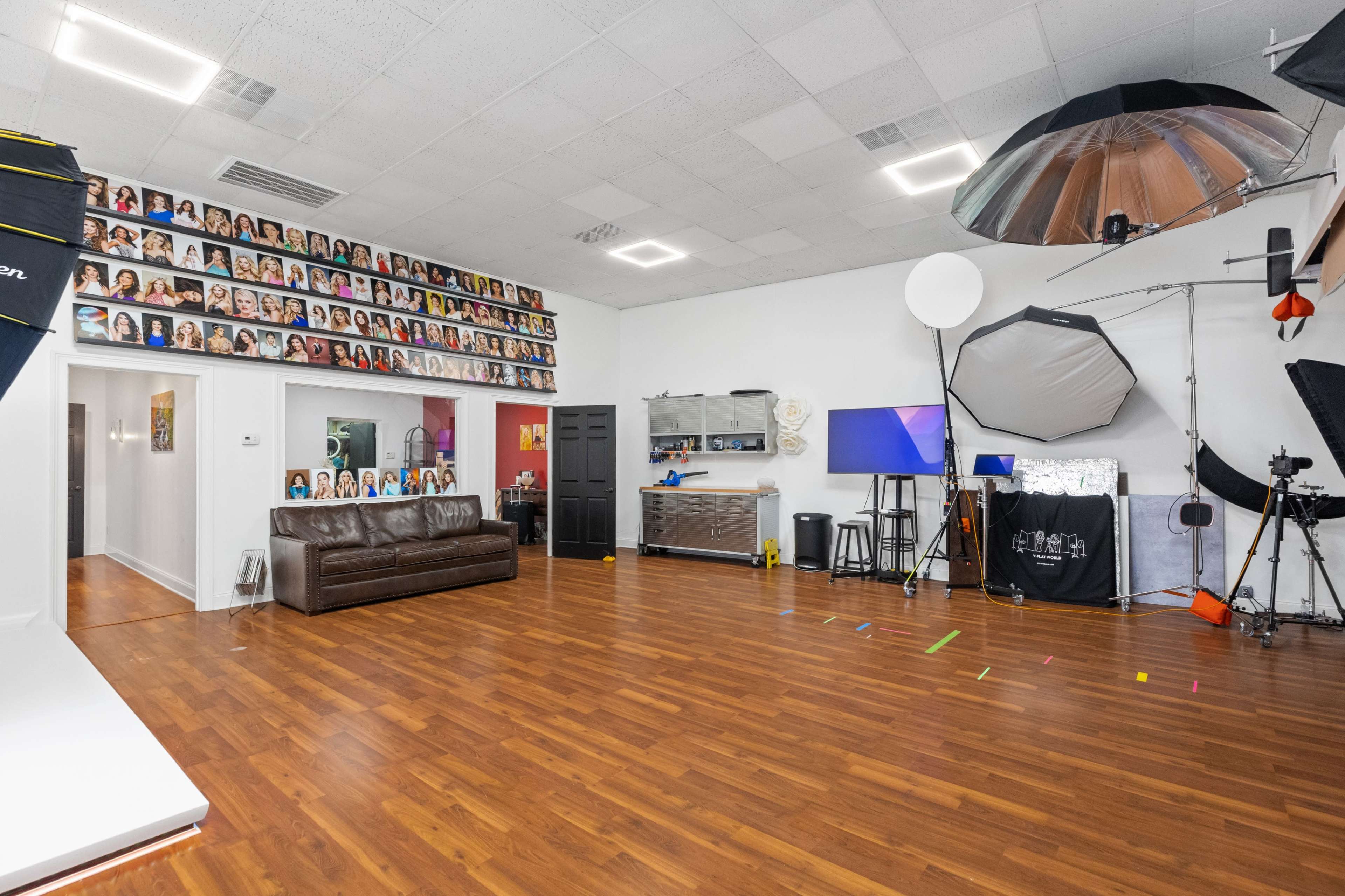 The image shows a well-lit photography studio featuring a brown couch, a wall adorned with numerous portrait photographs, and various lighting equipment set up for a photo shoot.