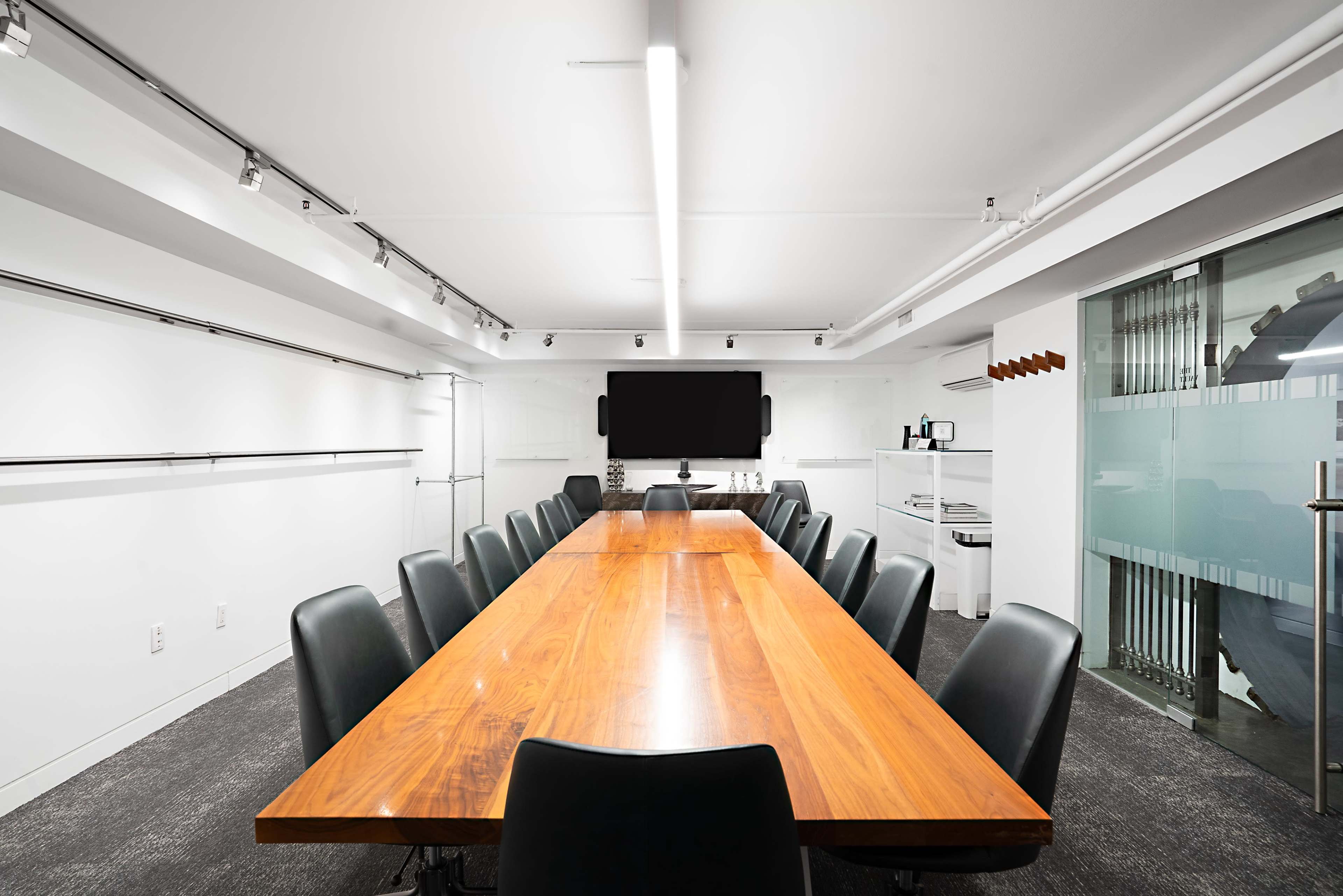 A long wooden conference table is surrounded by black chairs in a well-lit meeting room featuring a large screen on one wall.