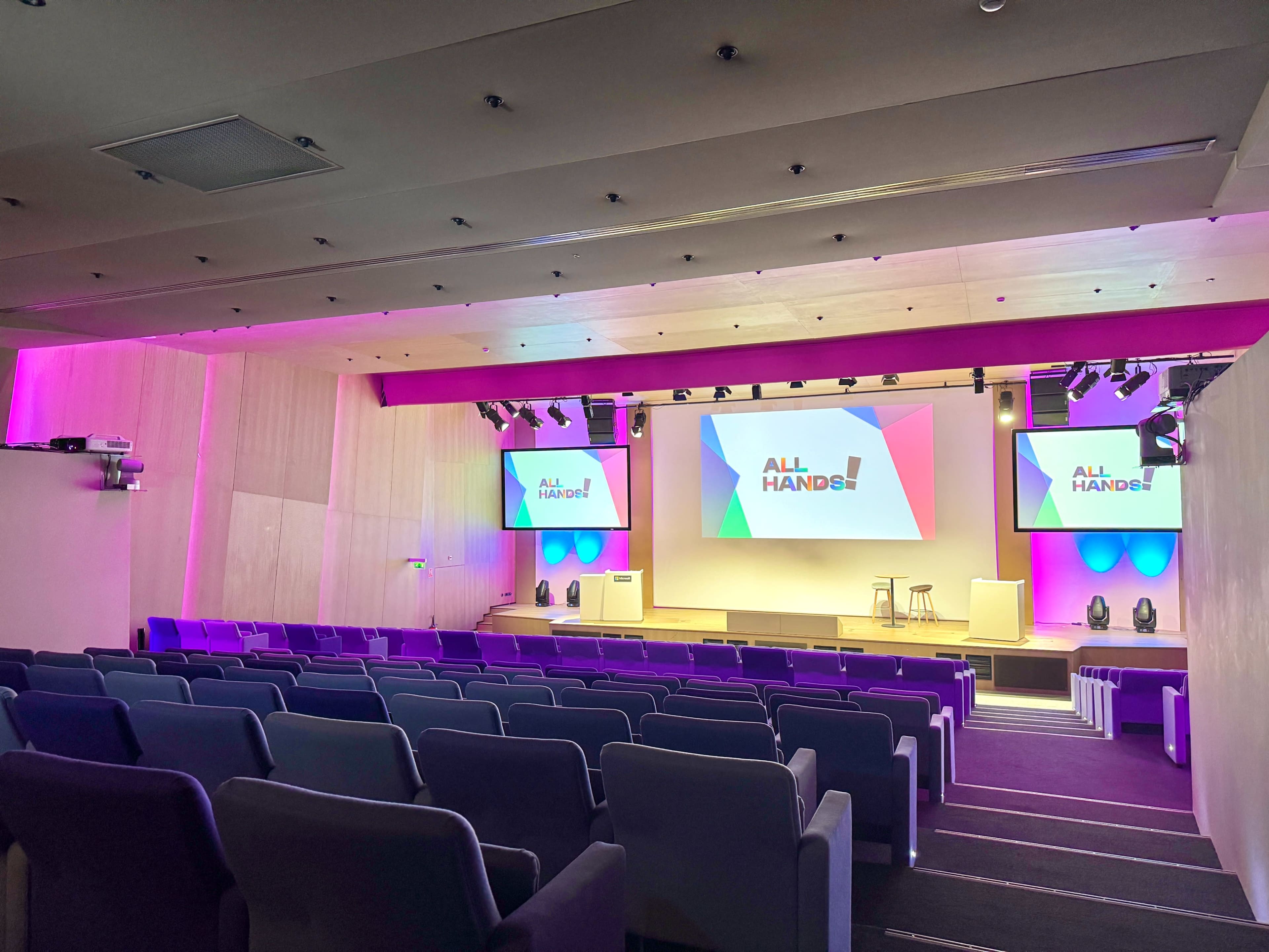 The image shows a modern conference room set up for a presentation, featuring rows of seats facing a stage with colorful screens displaying the text "ALL HANDS!"