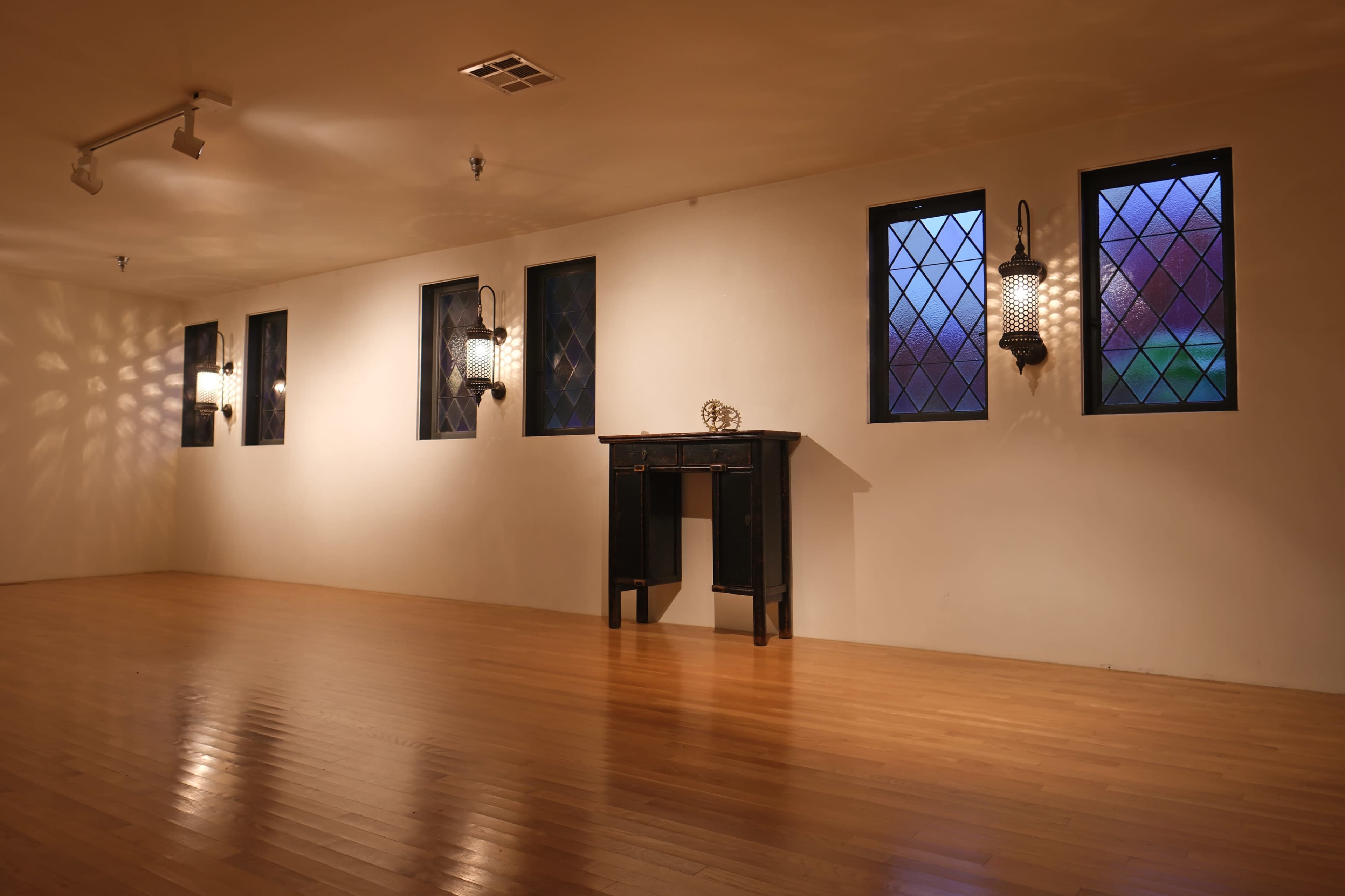 The image shows a spacious, empty room with hardwood flooring and wall-mounted lanterns, featuring diamond-patterned windows that allow soft light to enter.