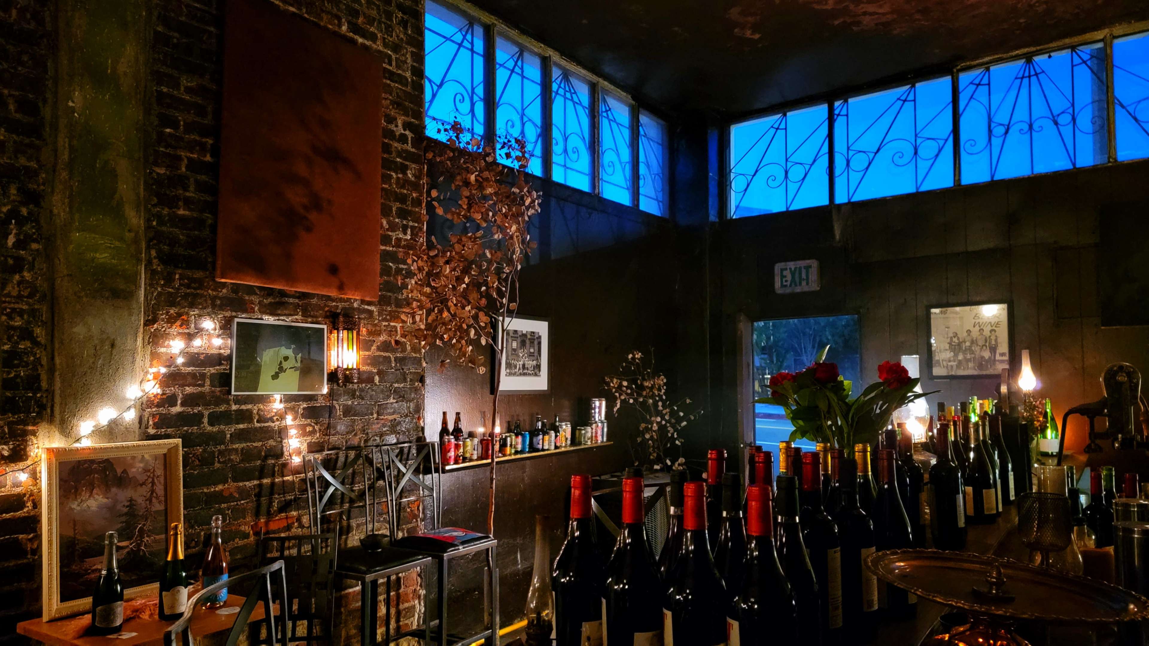The interior of a dimly lit wine bar features rows of wine bottles on a counter, with illuminated art and decorative elements against a brick wall and large windows.