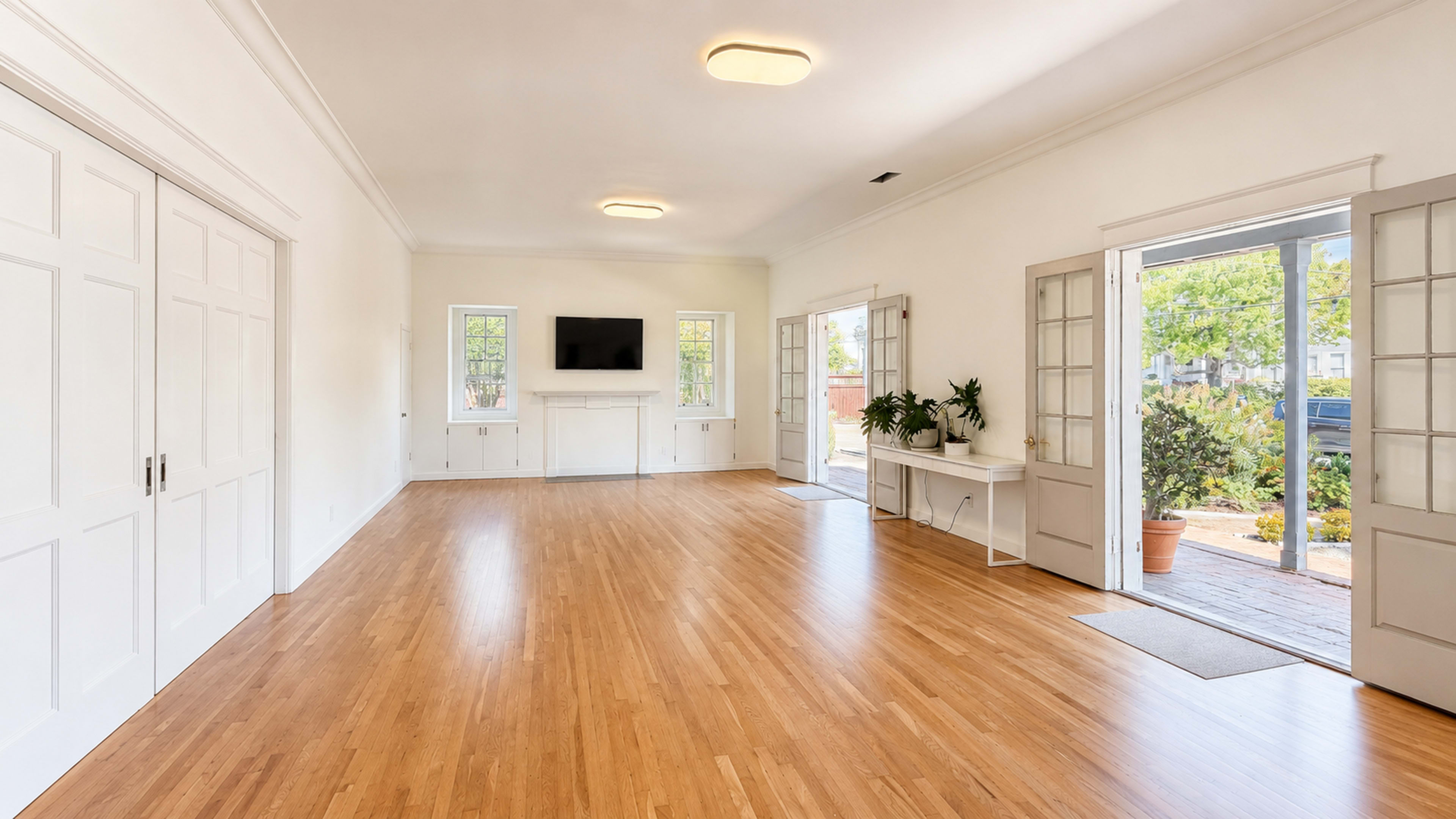 The image shows a spacious, empty room with hardwood floors, white walls, and large windows, leading to an outdoor area.