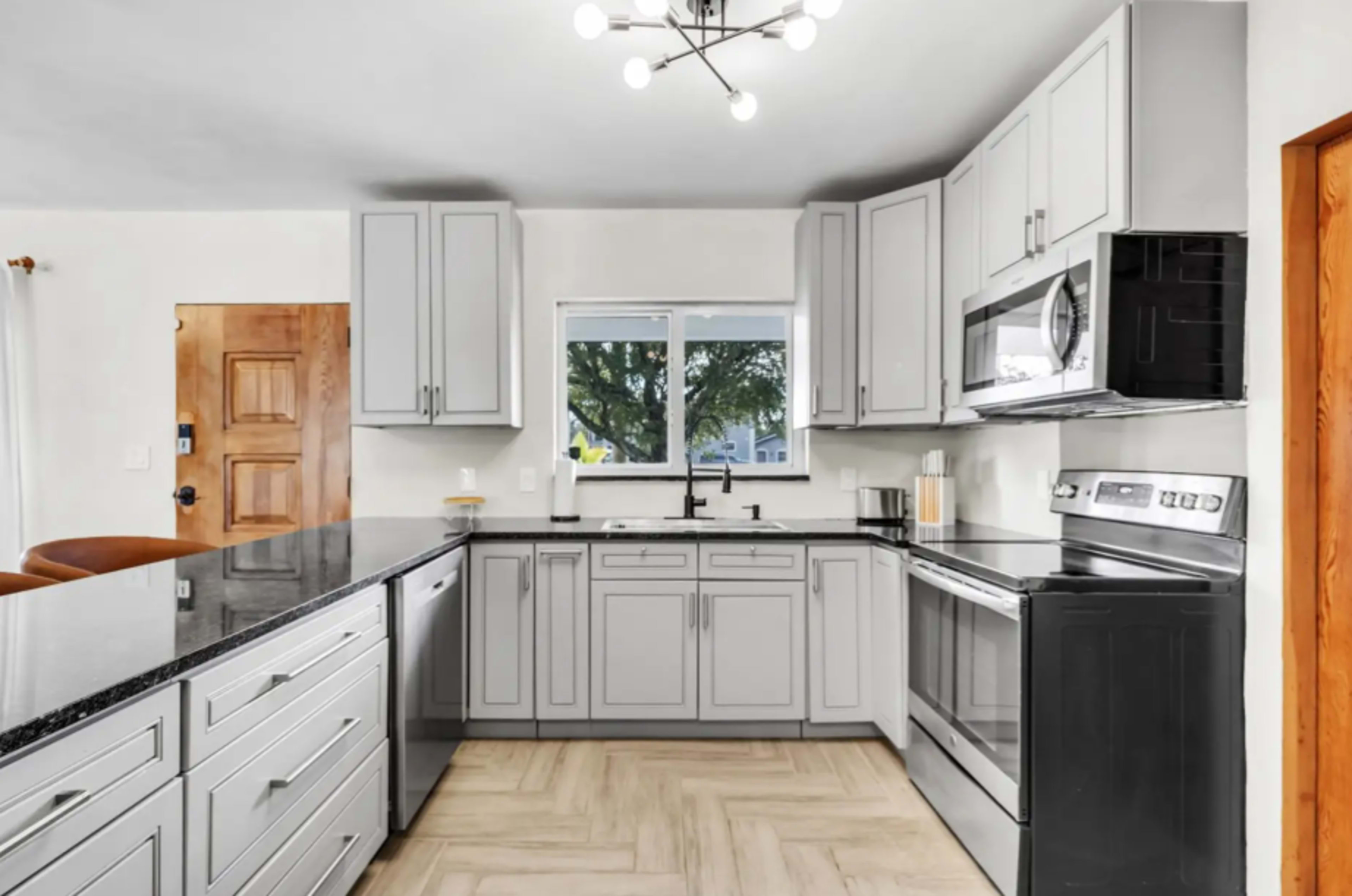 The image shows a modern kitchen featuring gray cabinets, a black countertop, stainless steel appliances, and a window above the sink.