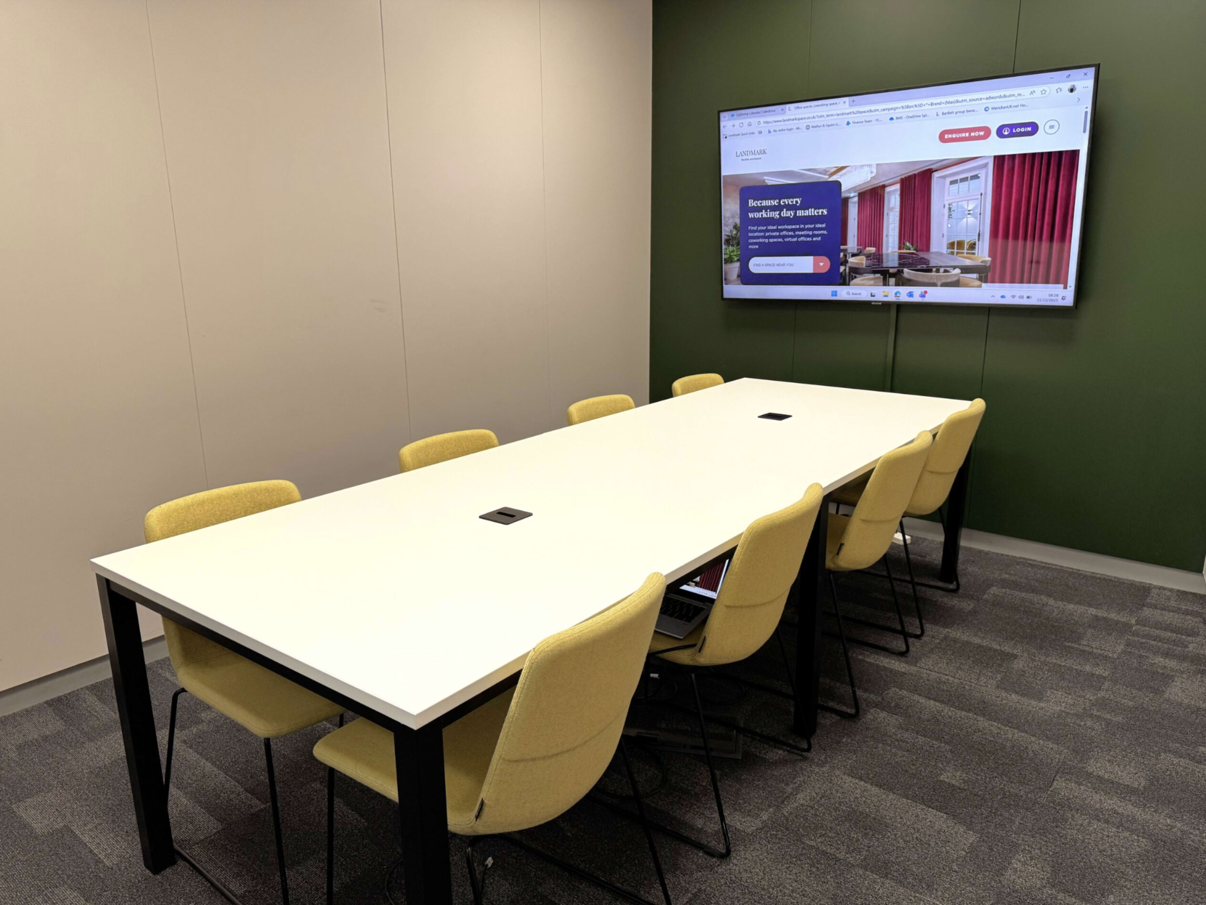 A long white table is surrounded by yellow chairs in a modern conference room, featuring a large screen displaying a website.