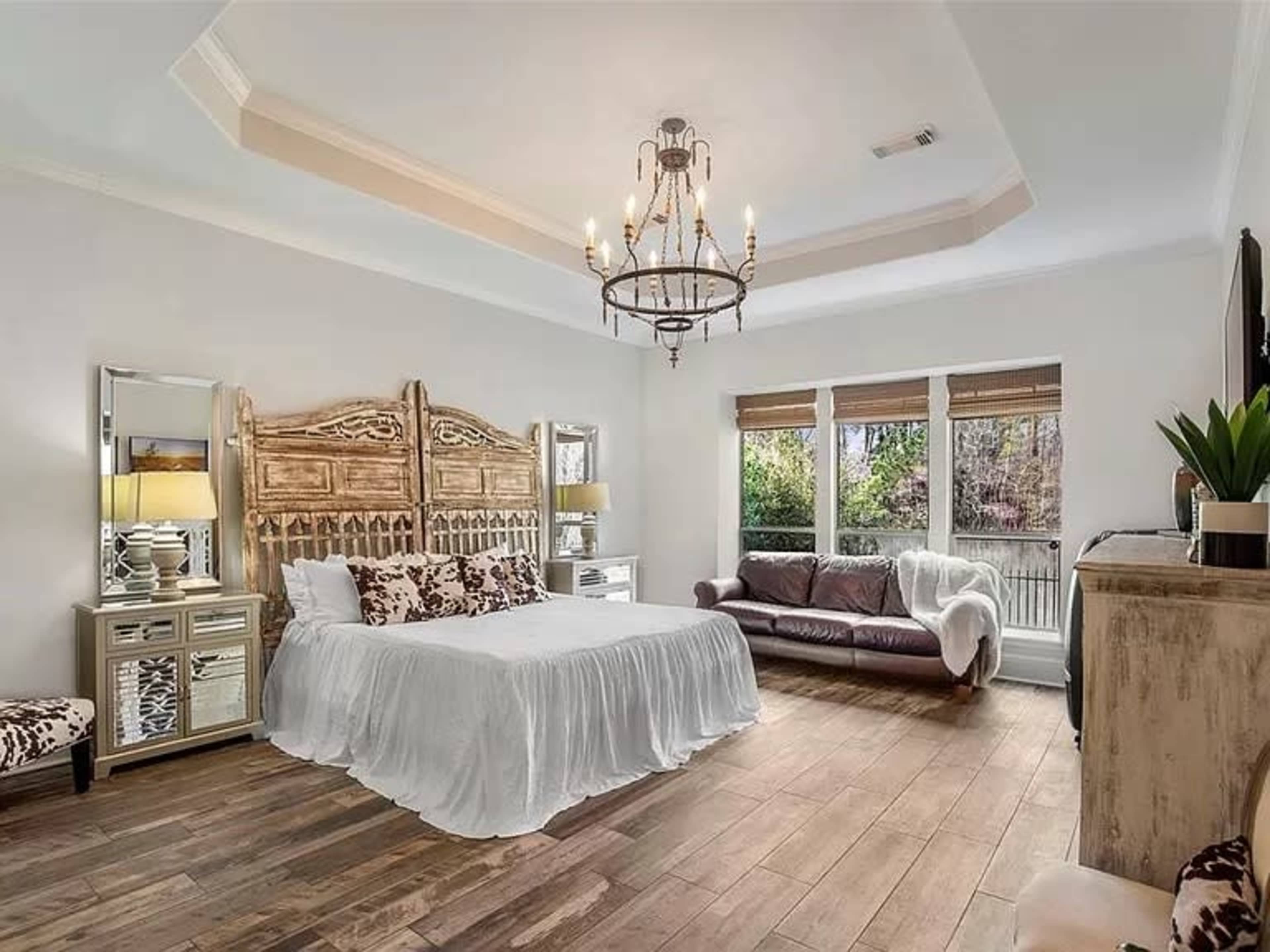 The image shows a spacious bedroom featuring a wooden bed frame, large windows with blinds, and a mix of light and dark furniture.