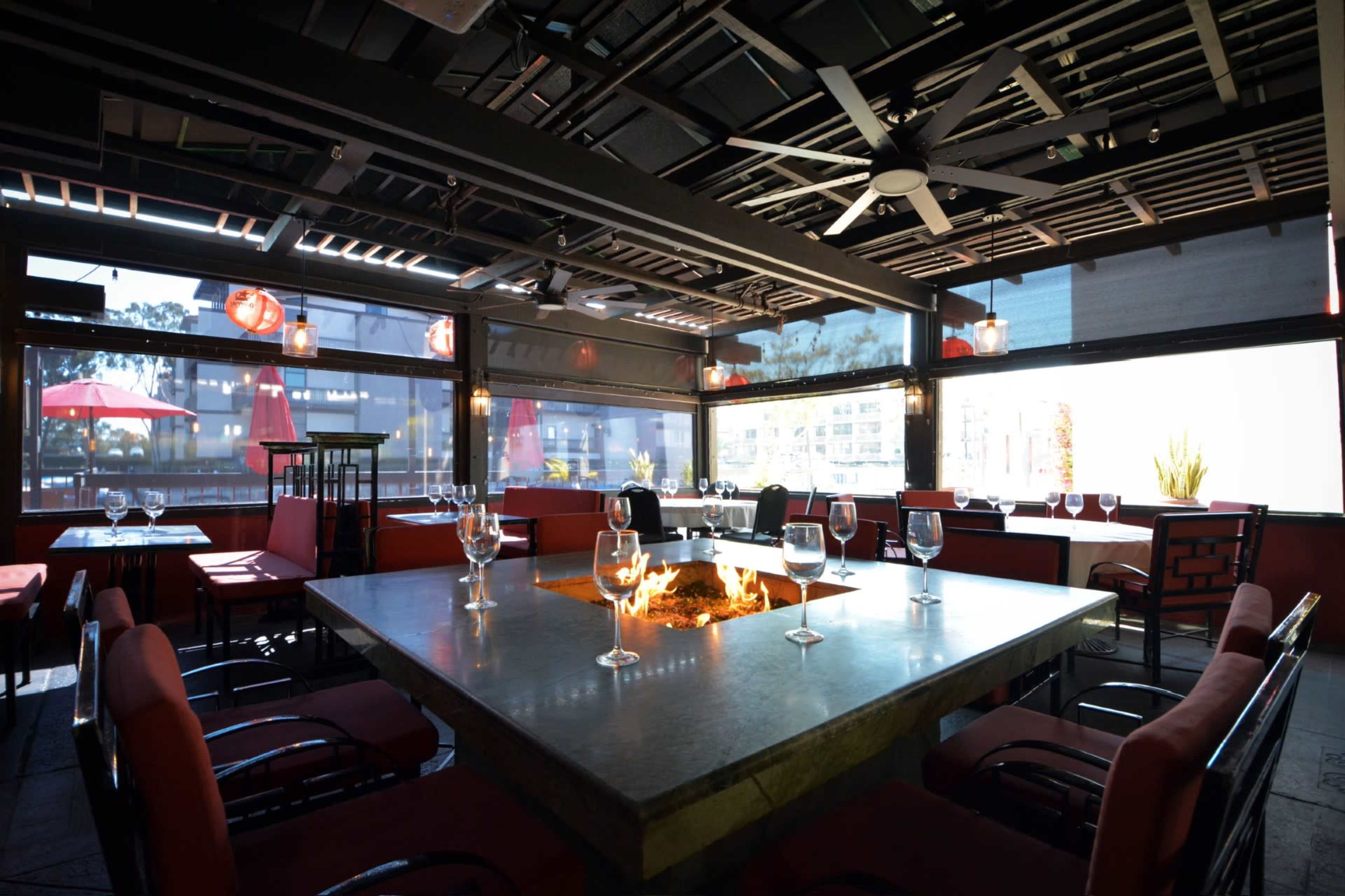 A modern restaurant interior with a large table surrounded by red chairs, featuring a fire pit at the center and large windows providing outdoor views.