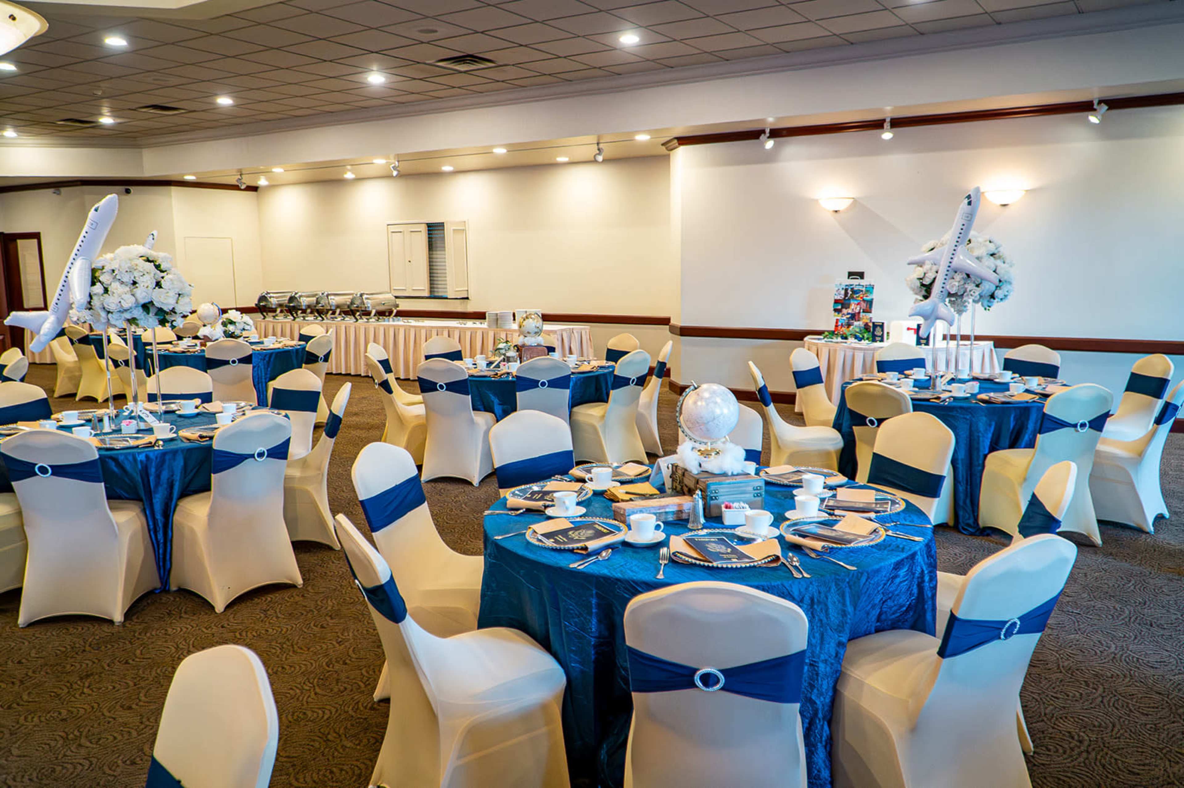 The image shows a banquet hall set up with round tables covered in blue tablecloths and adorned with white floral centerpieces.