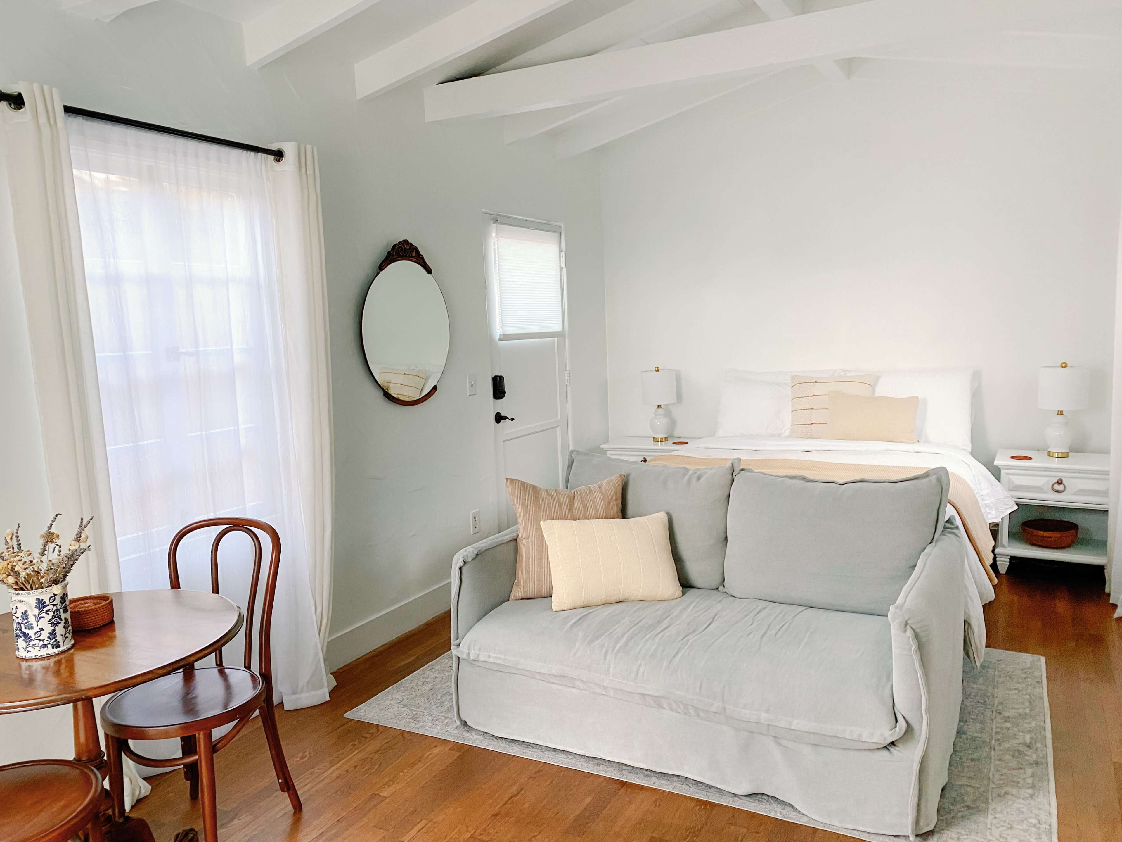 A bright, minimalistic interior featuring a bed, a light-colored sofa, a small dining table, and windows with sheer curtains.