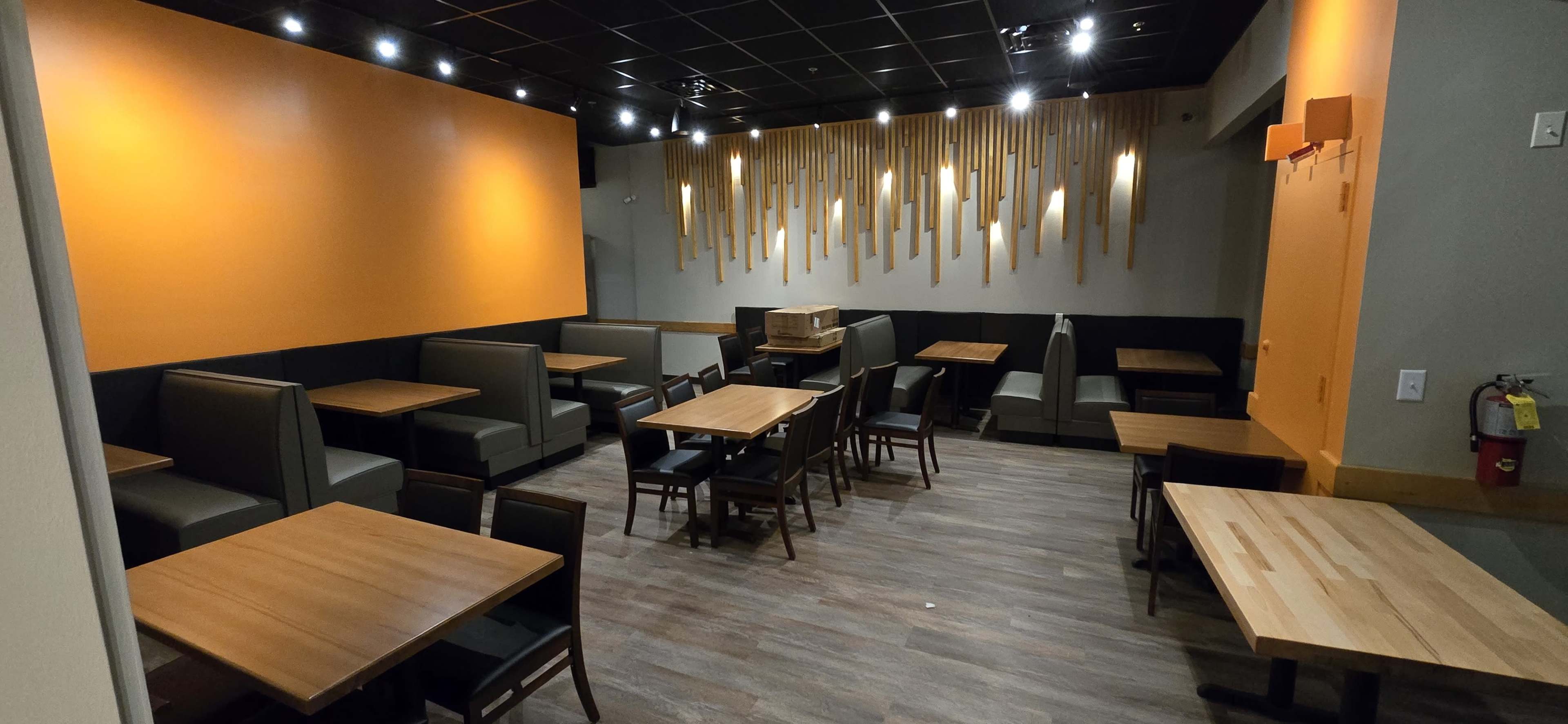 The image shows a modern restaurant interior with wooden tables and gray seating, featuring an accent wall in orange and decorative wooden elements hanging from the ceiling.