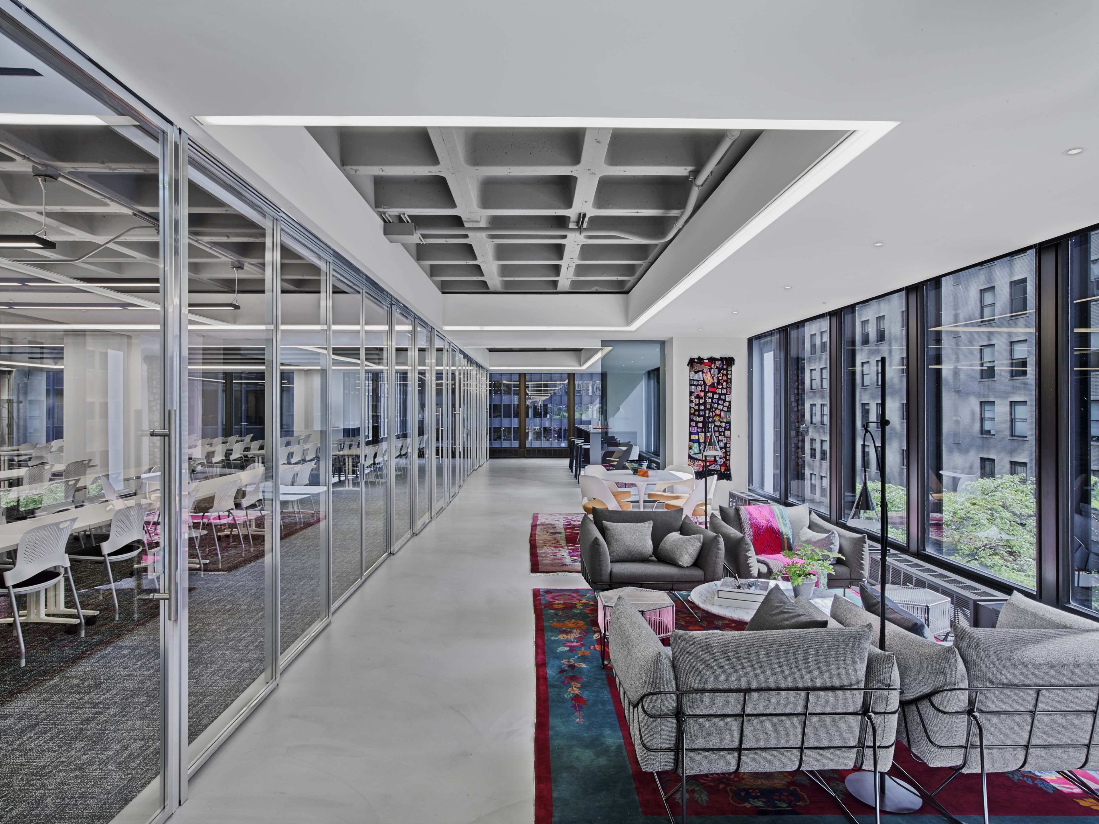 The image depicts a modern office space featuring a long corridor lined with glass partitions and contemporary seating areas decorated with colorful rugs and plants.