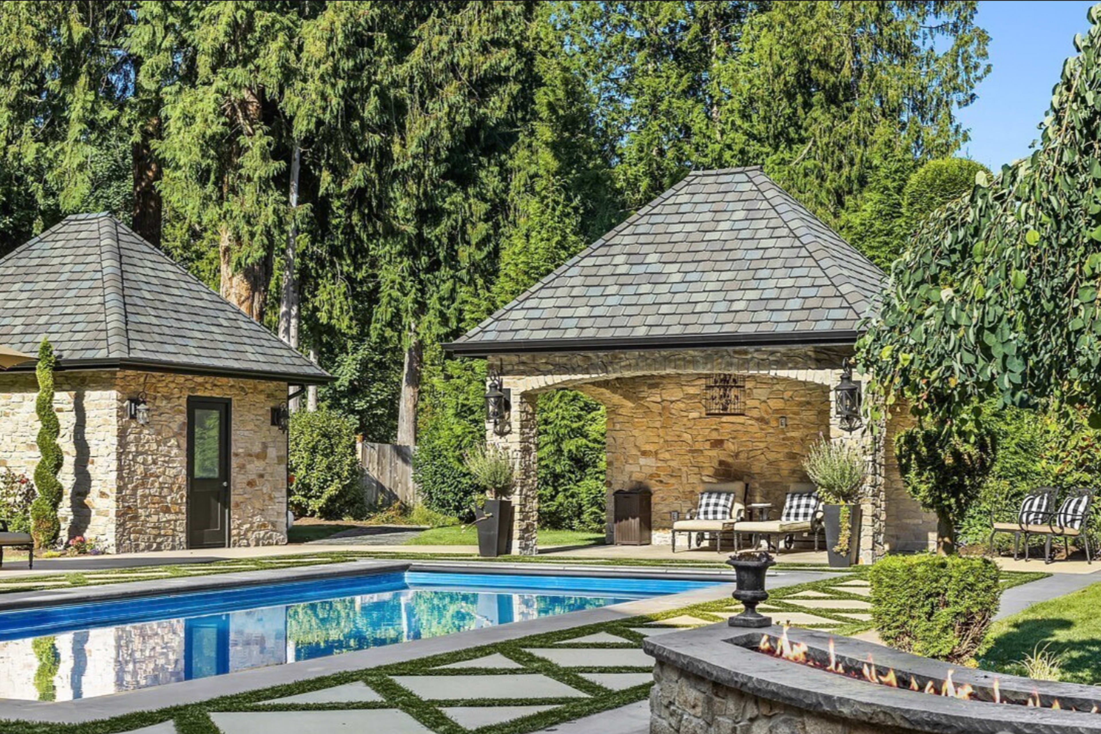 A well-maintained backyard features a stone patio with a swimming pool, surrounded by lush greenery and two identical gazebo structures.