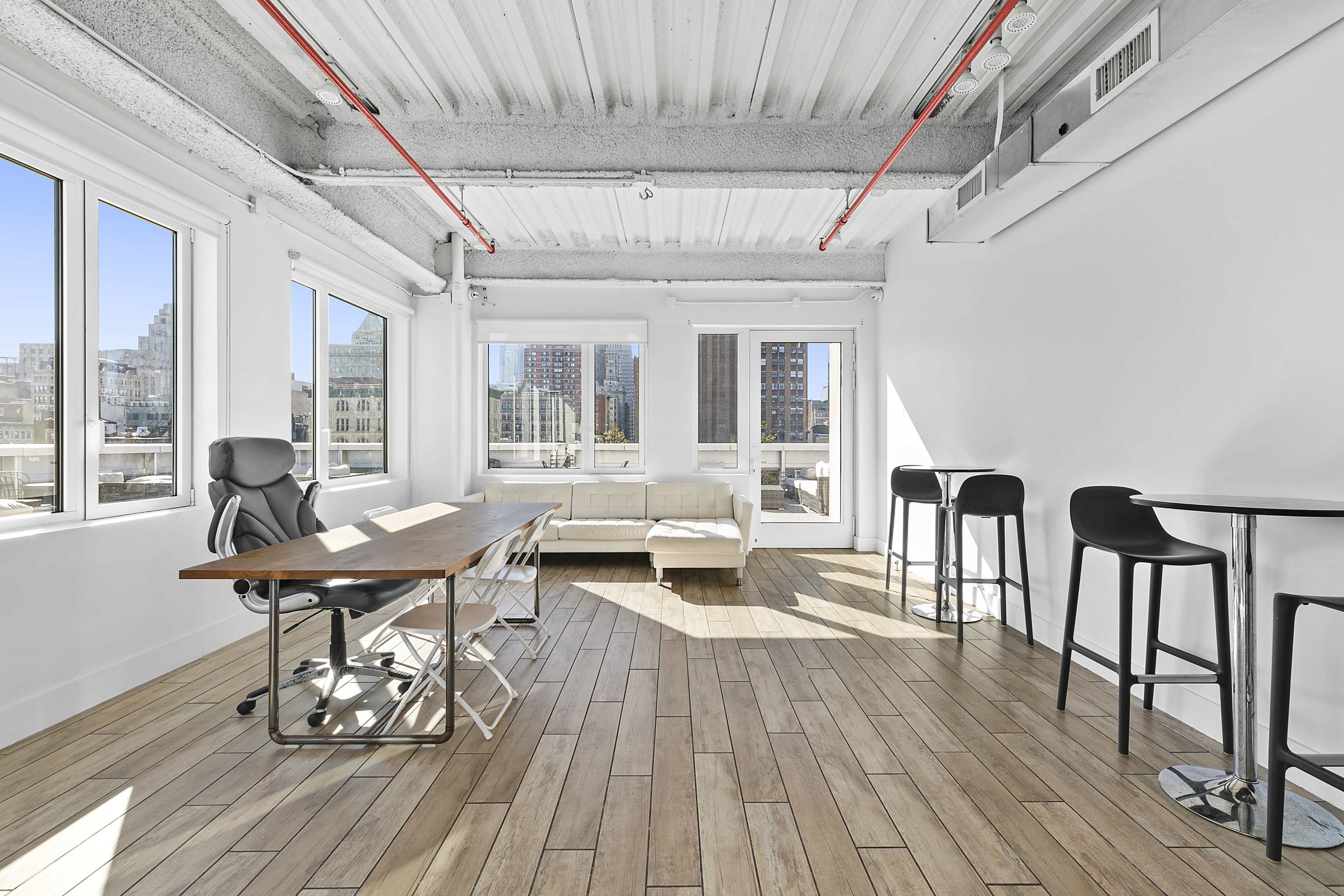 The image shows a bright, modern office space with a large wooden table, several chairs, a white sofa, and tall windows offering a view of the city.