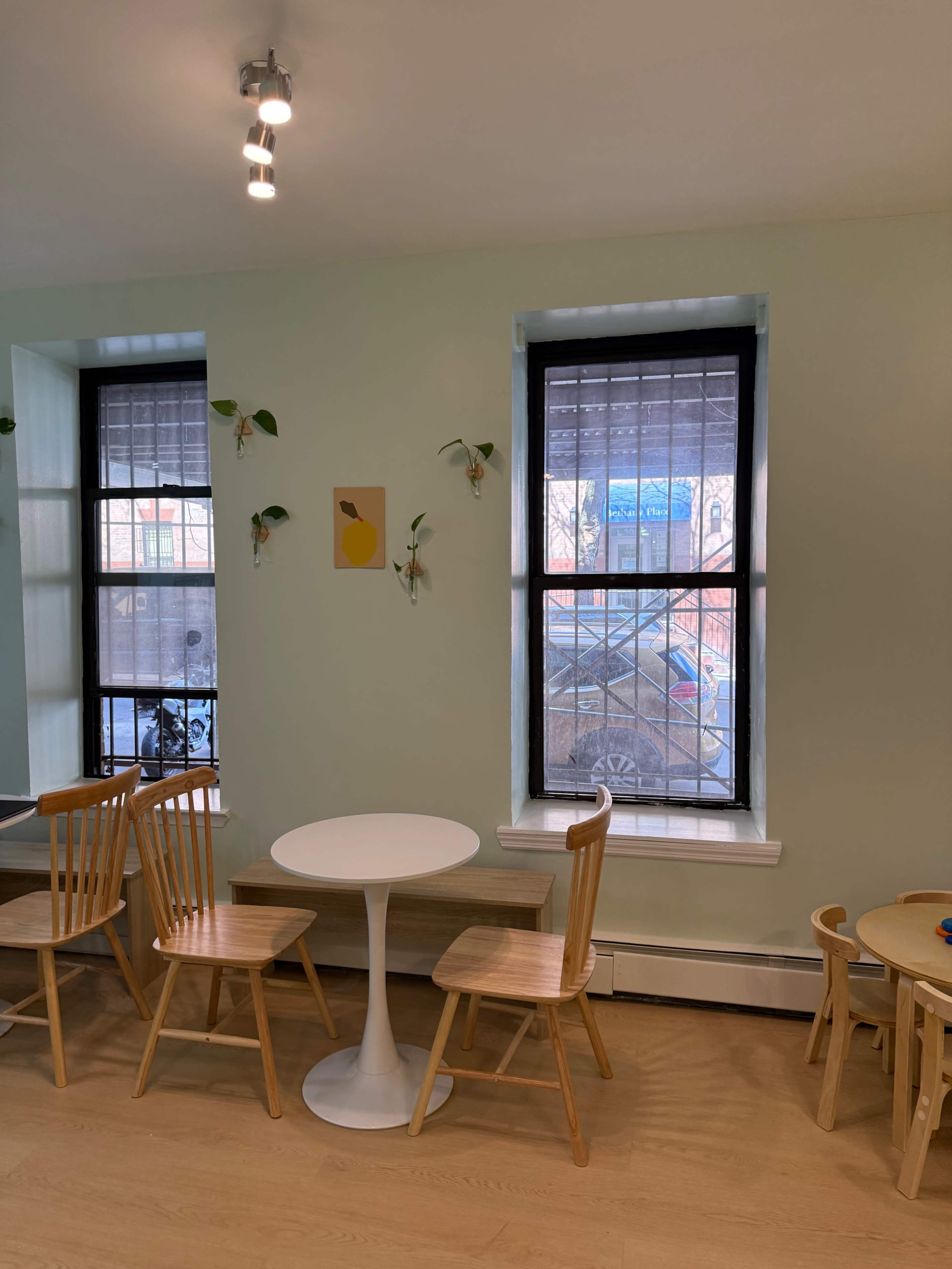 The image shows a light green room with two windows, a small round white table, and several wooden chairs arranged around it.