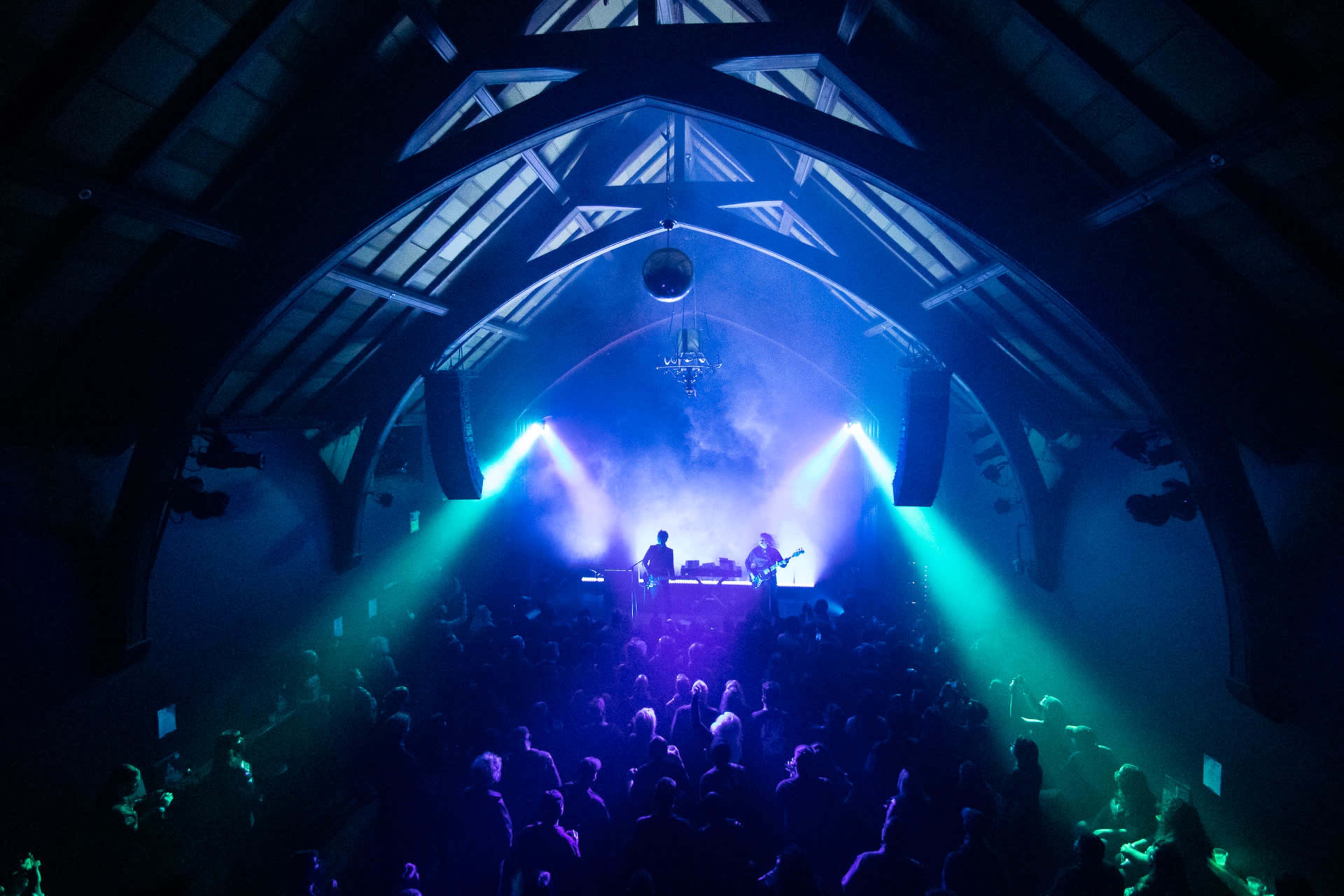 A large crowd gathers inside a dimly lit venue with a high, arched ceiling while a performer plays on stage, illuminated by colorful lights and a disco ball overhead.