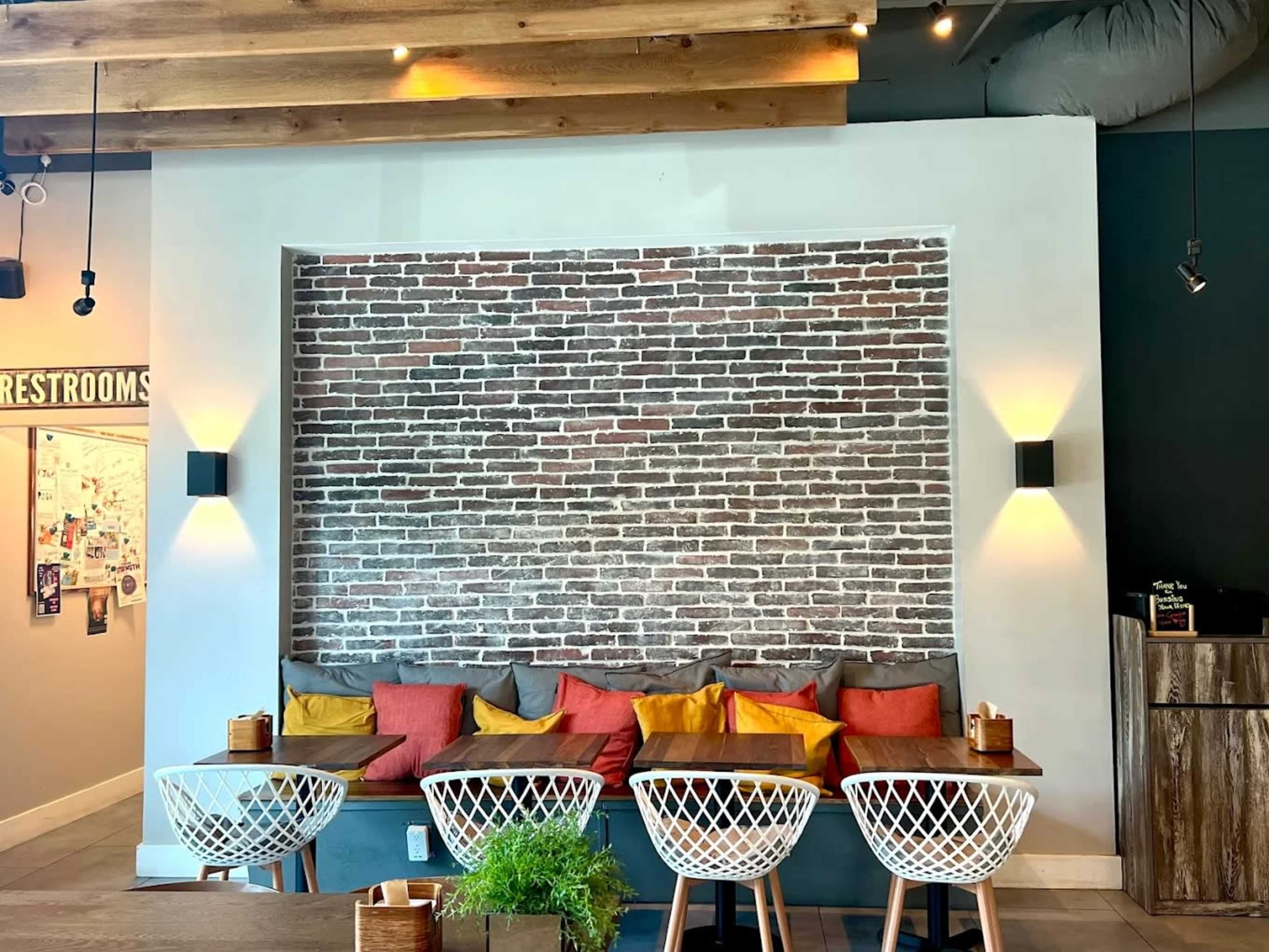 The image shows a cozy cafe interior featuring a brick wall, colorful cushions on a built-in seating area, and wooden tables with white wire chairs.