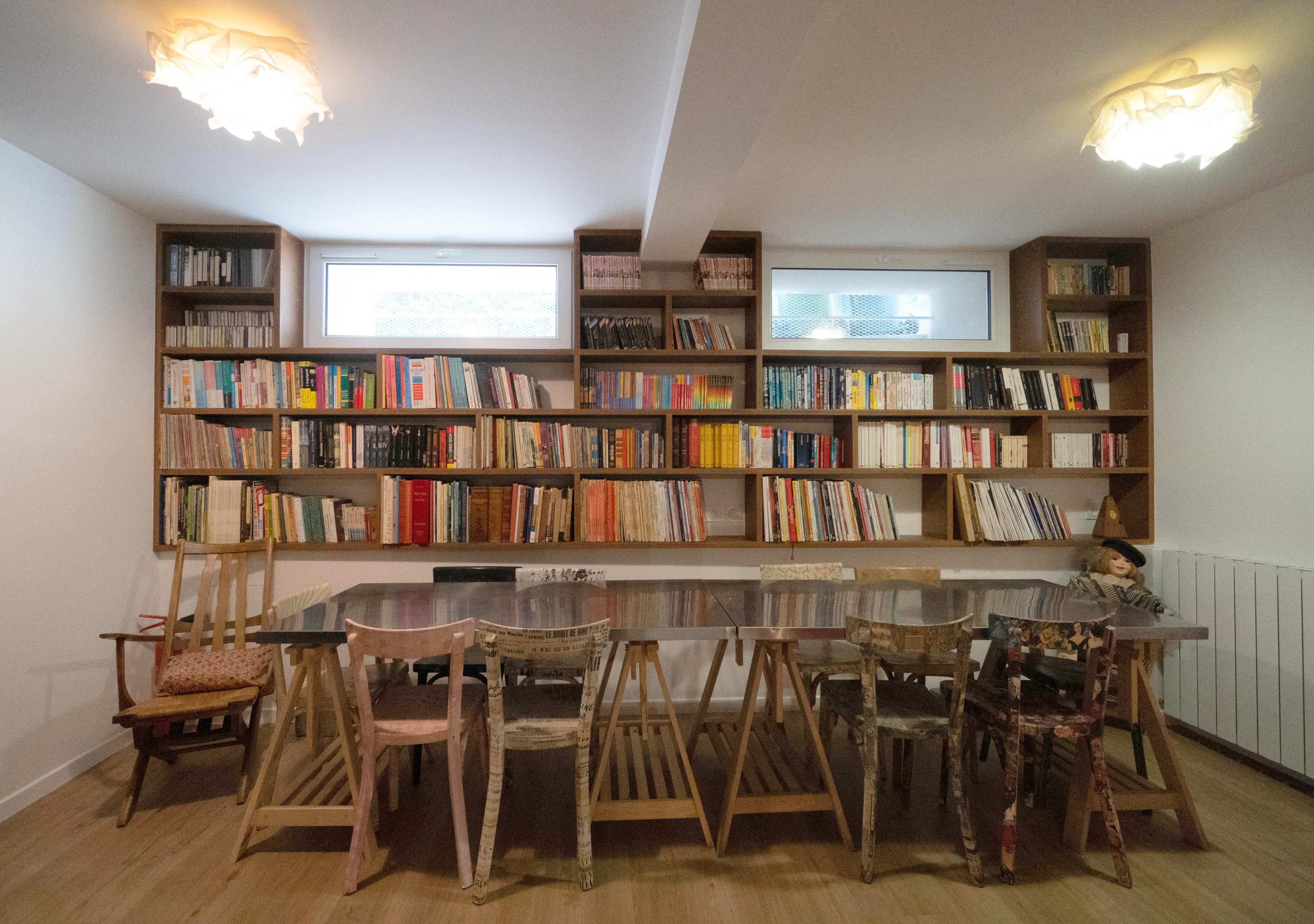 A spacious reading room features a long table surrounded by mismatched chairs, with bookshelves filled with various books lining the walls.