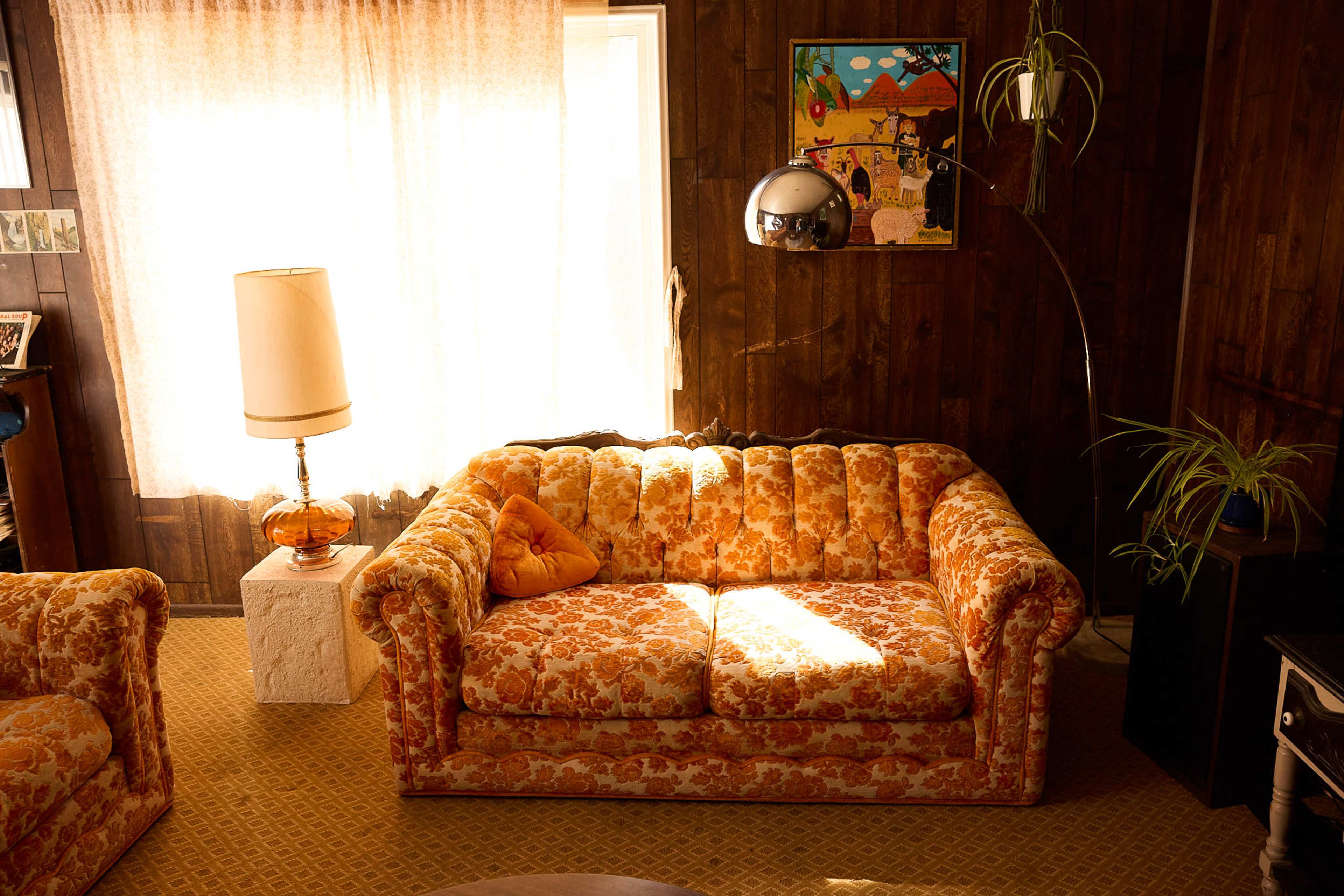 The image shows a brightly-lit room featuring a floral-patterned sofa, a floor lamp, and a decorative wall hanging.
