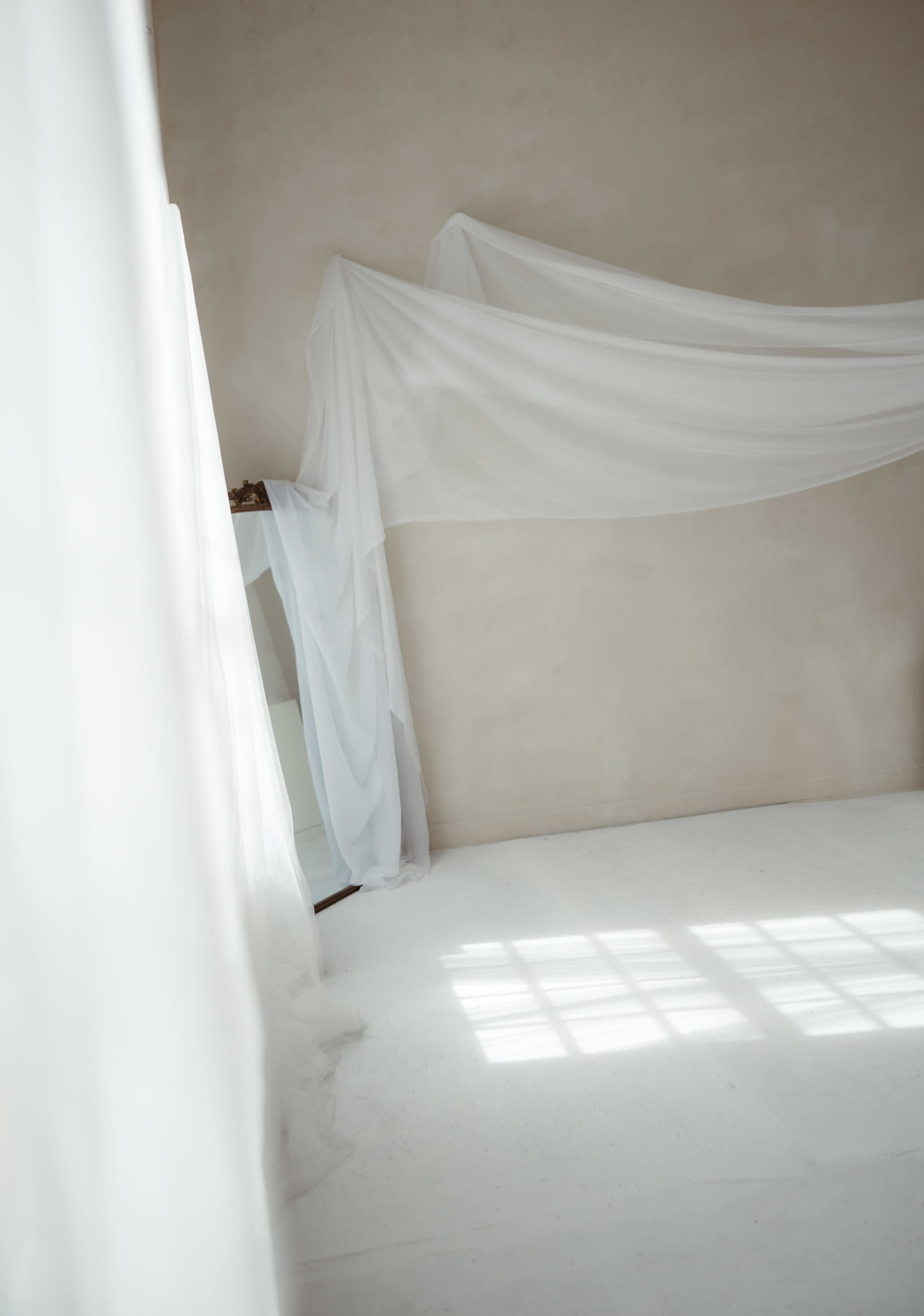 The image shows a minimalistic interior space with soft, draped white fabric and a large mirror against a light-colored wall.