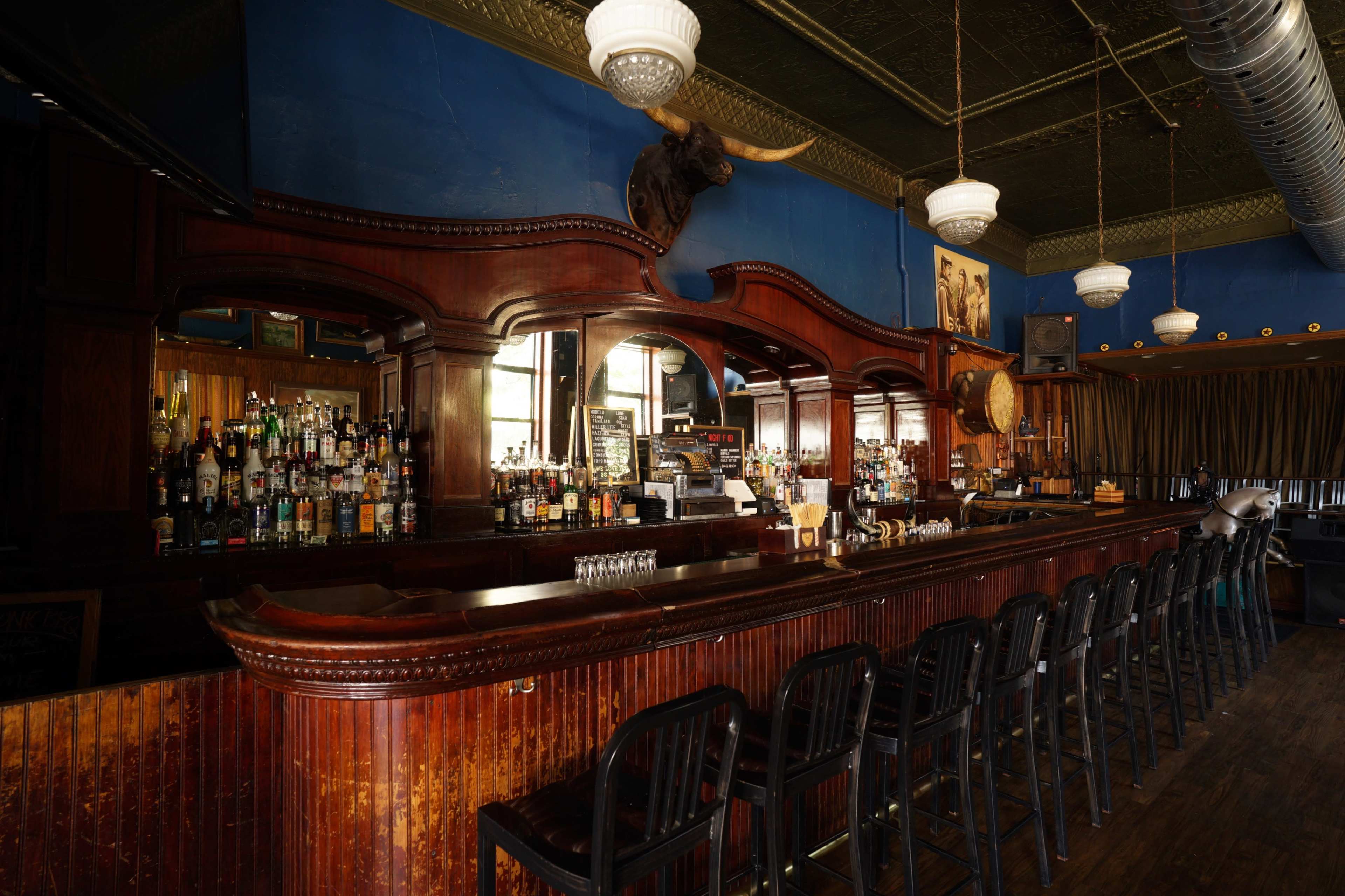 The image shows a wooden bar with high stools, a wide selection of bottles on shelves, and ornate decorations against a blue wall in a spacious room.